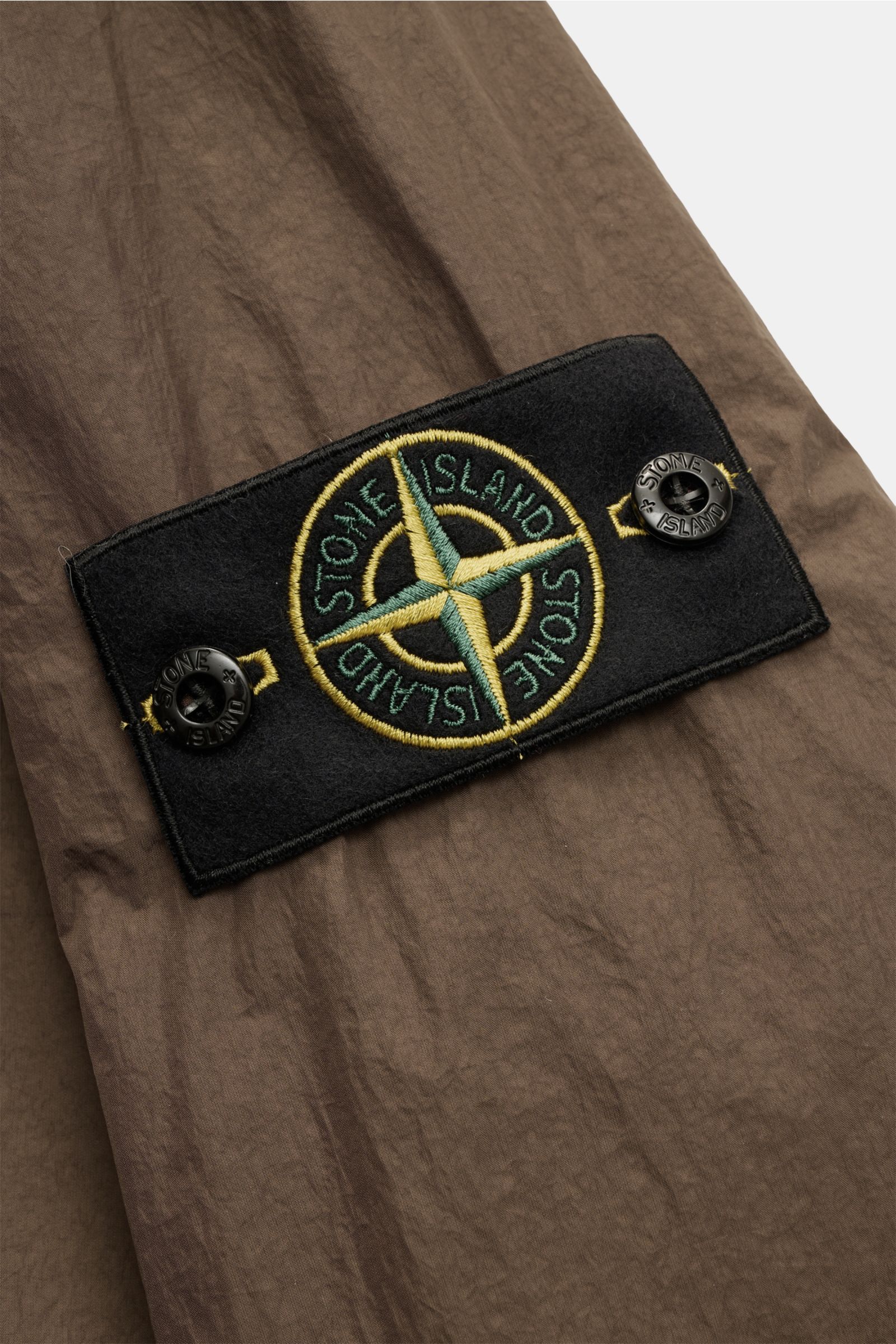Stone Island Jacke 'Garment Dyed Crincle Reps NY' braun, Nahaufnahme des schwarzen Kompass-Badges auf wasserabweisendem, windblockendem Material.