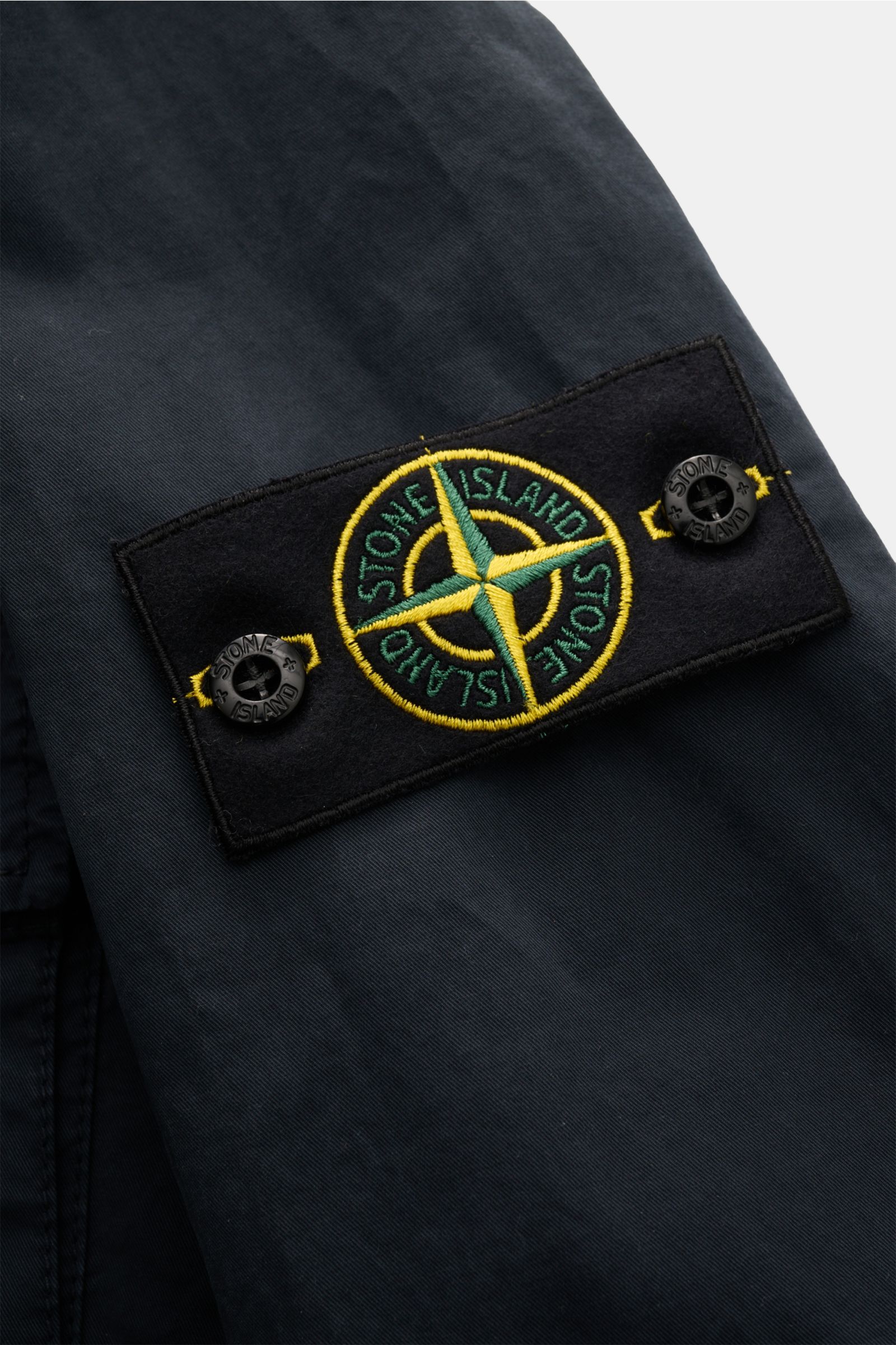 Close-up side view of Stone Island Blouson 'Stretch Cotton Twill-TC' navy showing the embroidered logo patch with buttons on elastic cotton fabric. Sportives Frühlings-Essential: Mit seinem leichten Baumwollmaterial wird der STONE ISLAND Blouson zum ideal