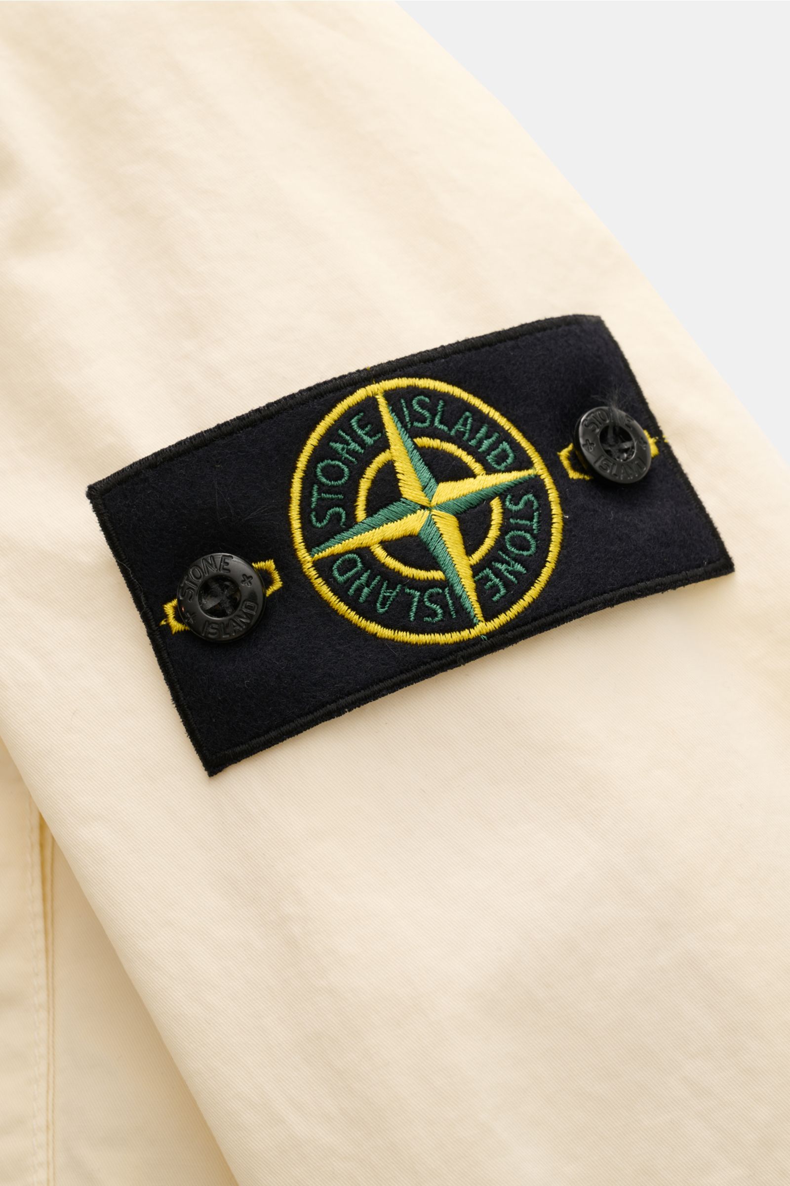 Nahaufnahme des schwarzen Stone Island Logos auf dem Ärmel des cremefarbenen Stone Island Blouson 'Stretch Cotton Twill-TC', sportiv aus elastischer Baumwolle, Slim Fit, mit weichem Griff, robustem Zwei-Wege-Reißverschluss, Kapuze mit Tunnelzug, aufgesetz