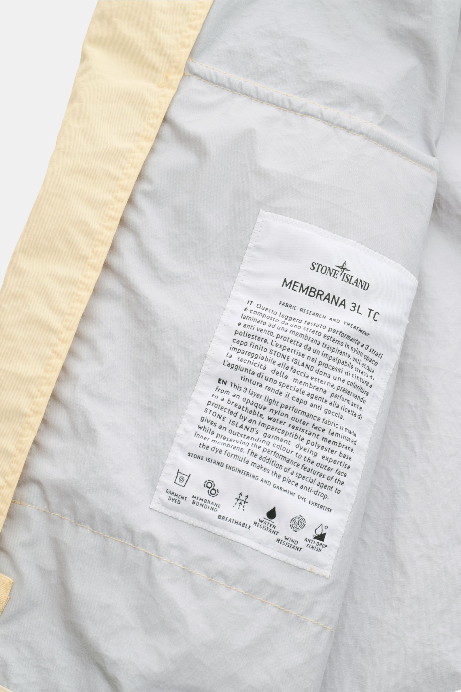 Nahaufnahme der Innenseite der Stone Island Jacke 'Membrana' creme mit weißem Label, das das dreilagige, wind- und wasserabweisende Funktionsgewebe beschreibt.