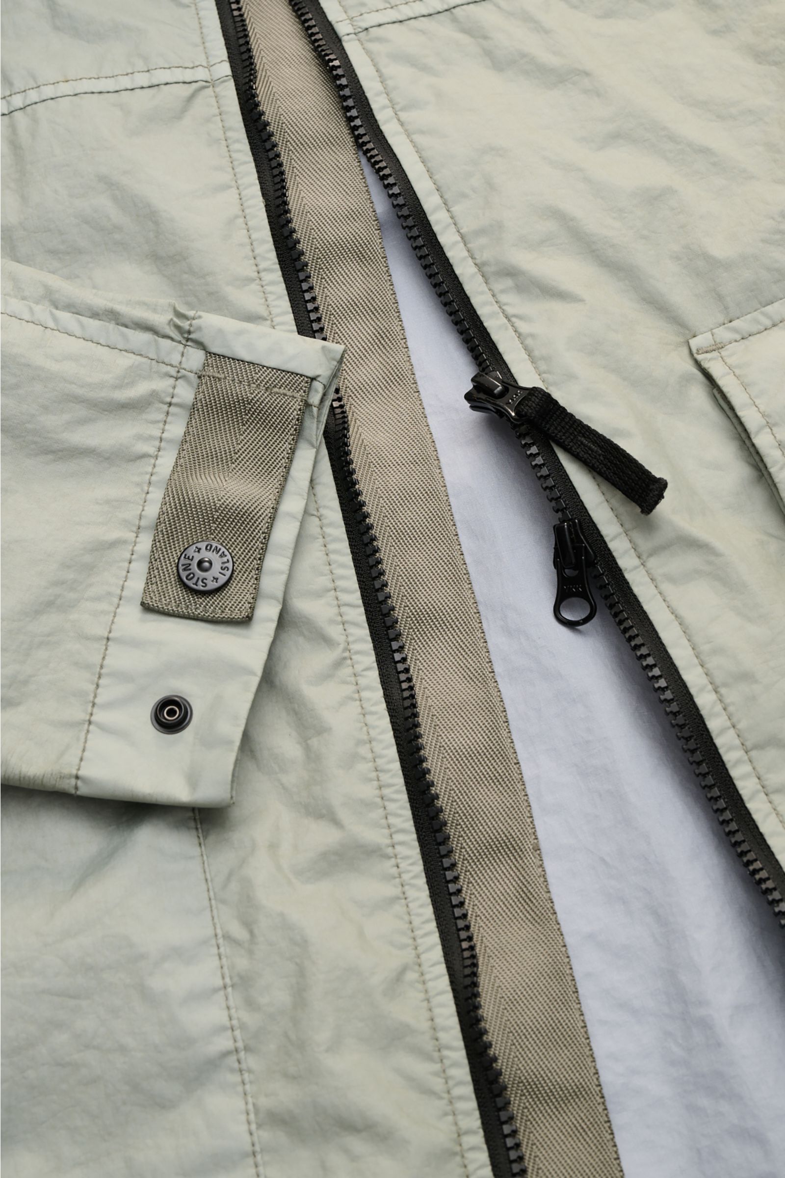Close-up view of Stone Island Jacket 'Membrana' sage showing the zipper, cuff with button, and lightweight textured fabric details. Function meets style: The triple-layer functional fabric of this STONE ISLAND jacket is laminated with a breathable, water 