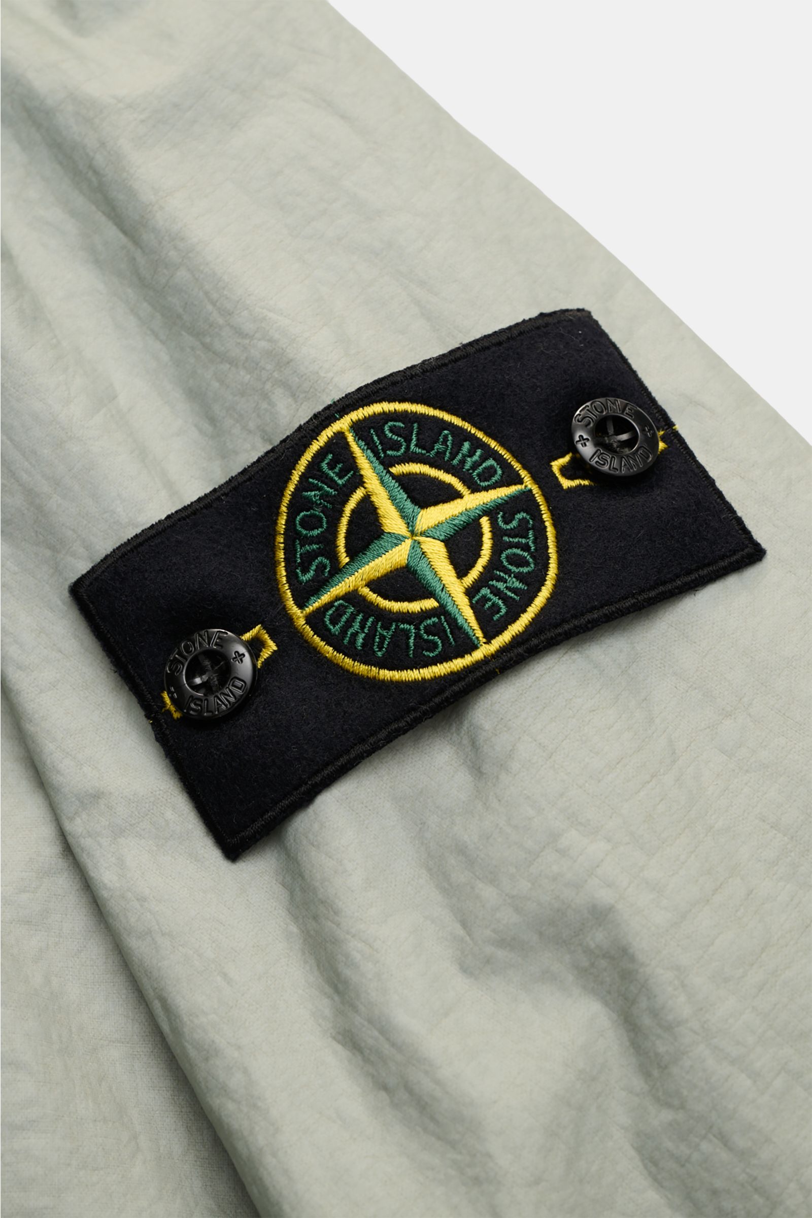 Close-up of the Stone Island Jacket 'Membrana' sage showing the embroidered compass badge on the upper sleeve from a top side angle, highlighting its lightweight, wind and water repellent fabric and unconstructed design. Function meets style: The triple-l