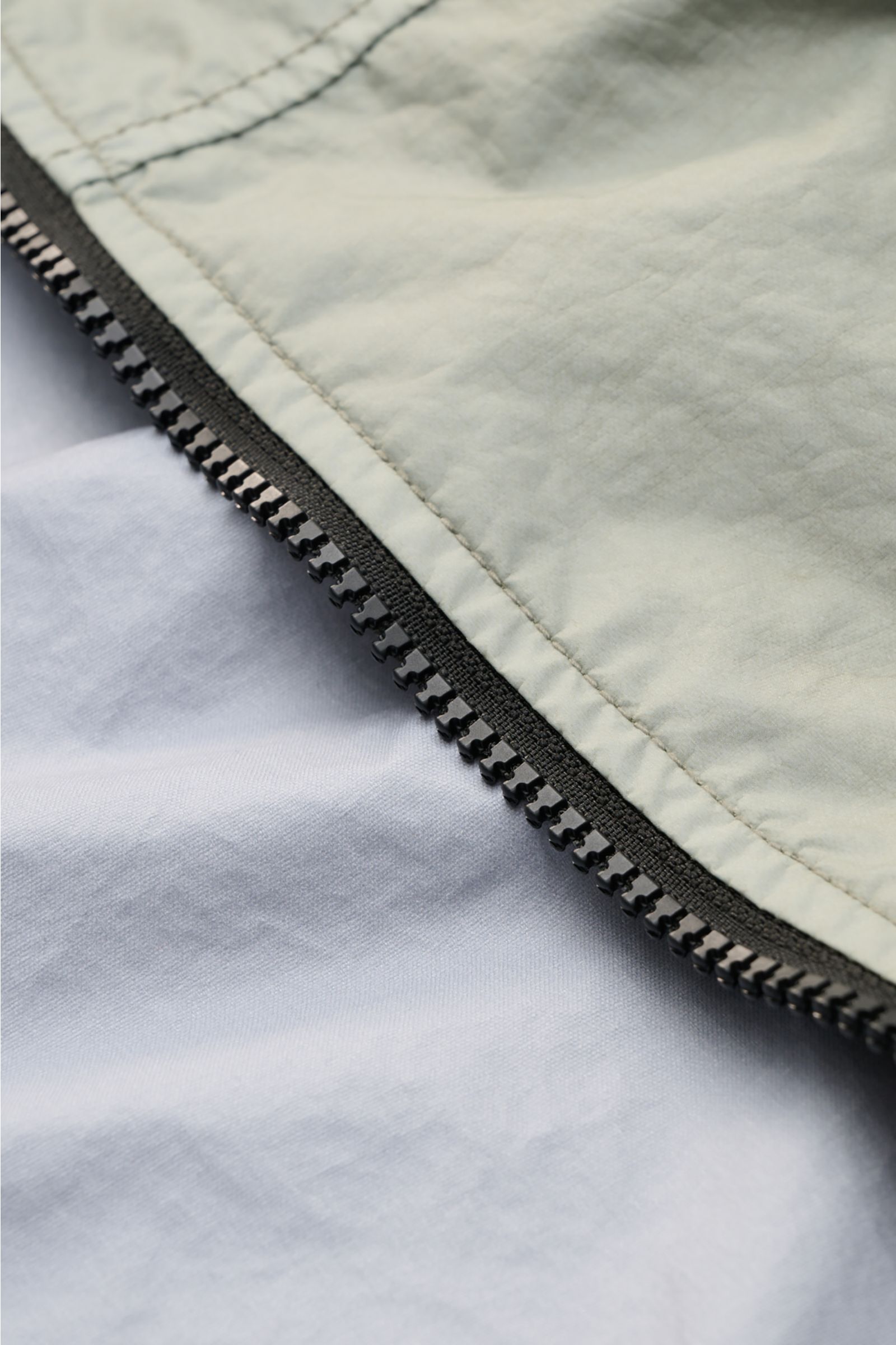 Close-up angled view of the zipper and fabric of the Stone Island Jacket 'Membrana' sage, highlighting its lightweight, wind and water repellent microfibre material. Function meets style: The triple-layer functional fabric of this STONE ISLAND jacket is l