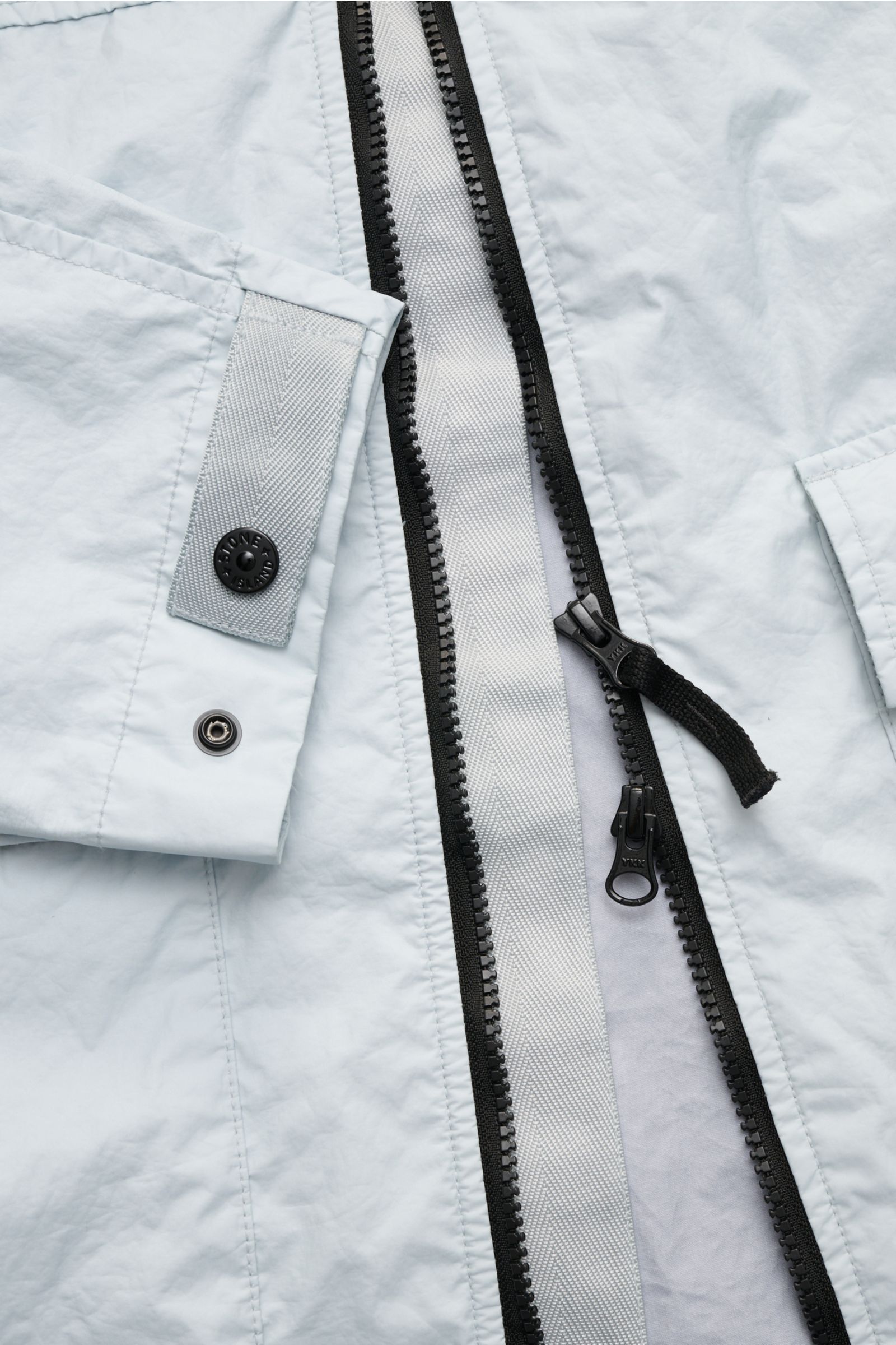 Light gray Stone Island Jacke 'Membrana' photographed close-up from above, showing the two-way black zipper, cuff with branded button, and smooth lightweight microfiber fabric with water-repellent membrane. Funktion trifft Style: Dreilagiges, leichtes Fun