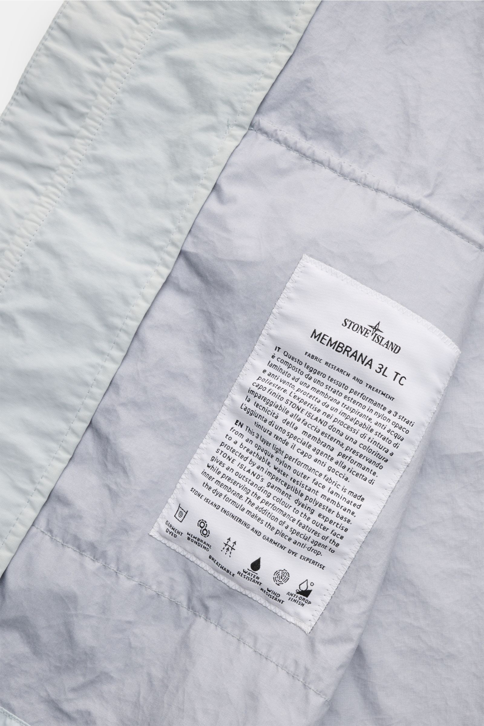 Close-up inside view of Stone Island Jacke 'Membrana' hellgrau showing fabric label detailing breathable, water-resistant membrane and three-layer lightweight microfiber design.