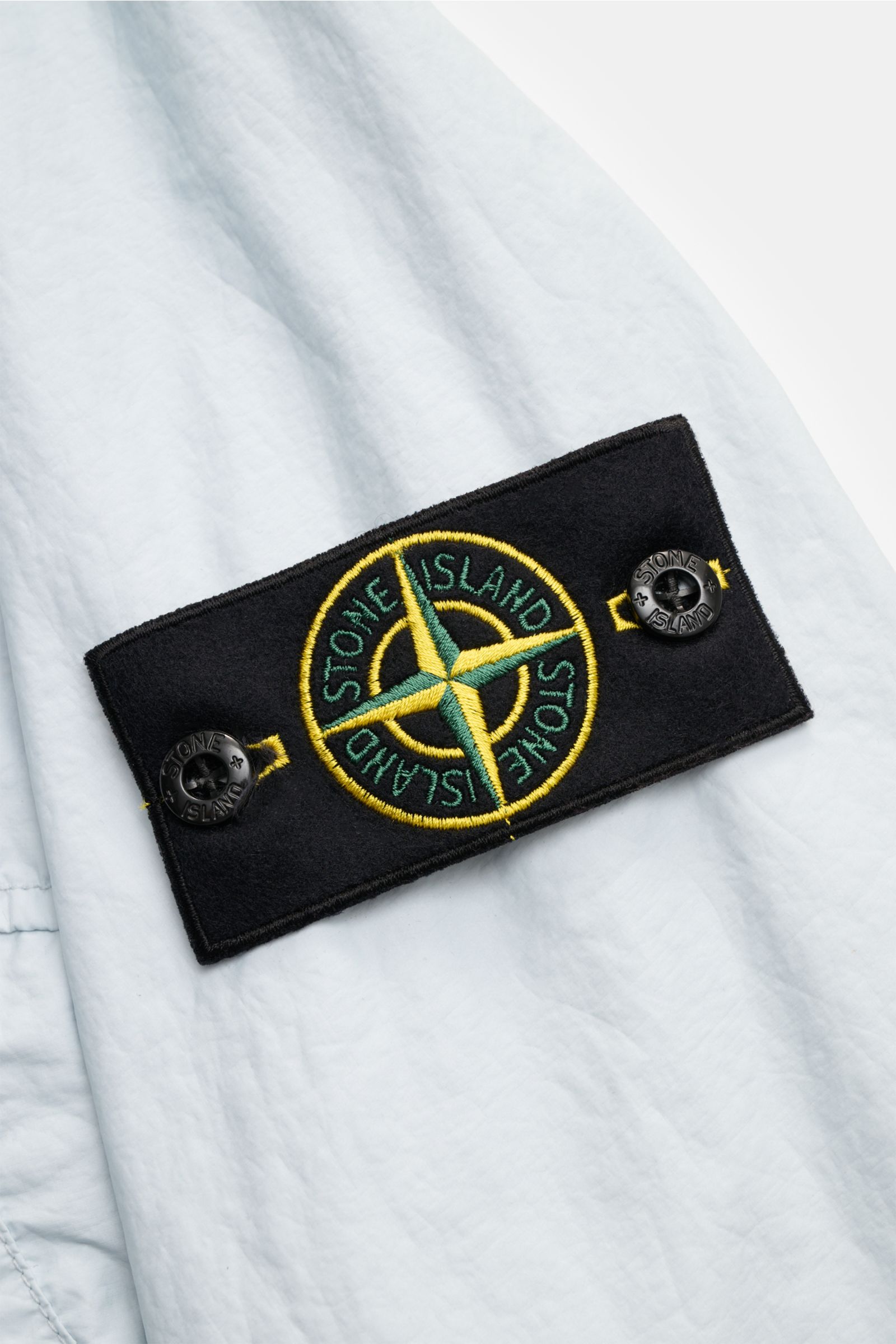 Close-up of the black compass badge on the sleeve of the light gray Stone Island Jacke 'Membrana' photographed from a side angle, showing lightweight, wind- and water-repellent microfiber fabric.