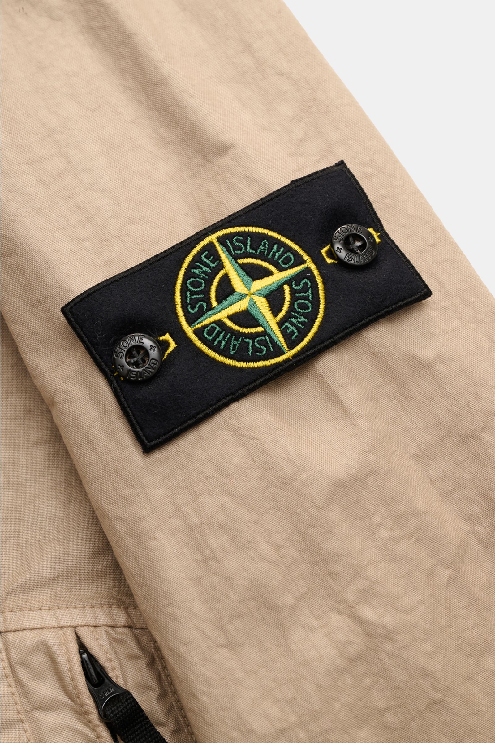 Close-up angled view of Stone Island Jacke 'Organic-Look Nylon Panama-TC' beige showing black compass badge with yellow and green embroidery on sleeve, highlighting high-quality microfiber, dense canvas, water-repellent fabric, slim fit, robust two-way zi