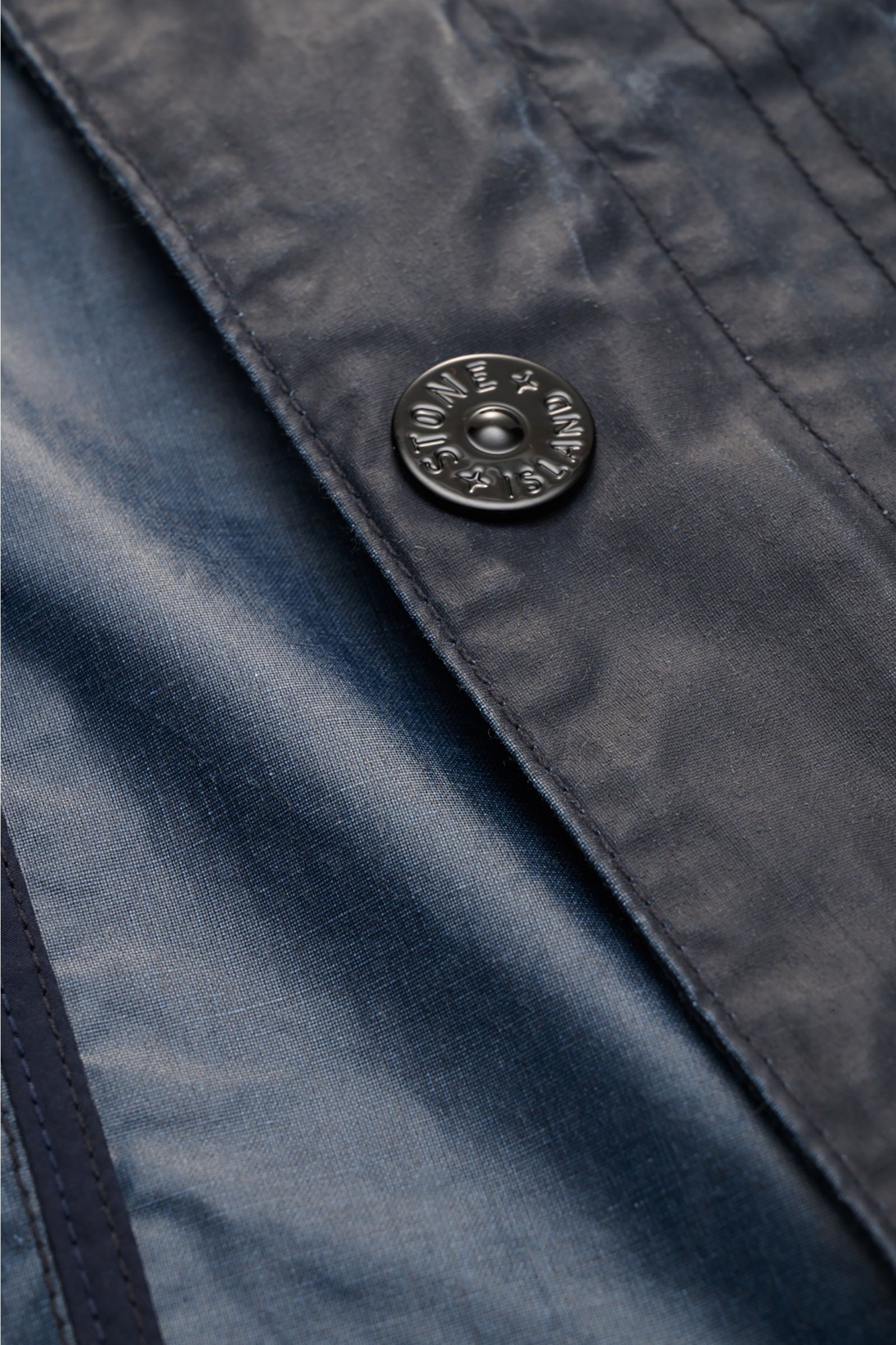 Close-up front detail of Stone Island Waxed jacket 'Waxed Pigment Cotton Tela' navy, showing water-repellent waxed cotton fabric and branded snap button.

The waxed jacket 'Waxed Pigment Cotton Tela' by STONE ISLAND features the expected level of innov