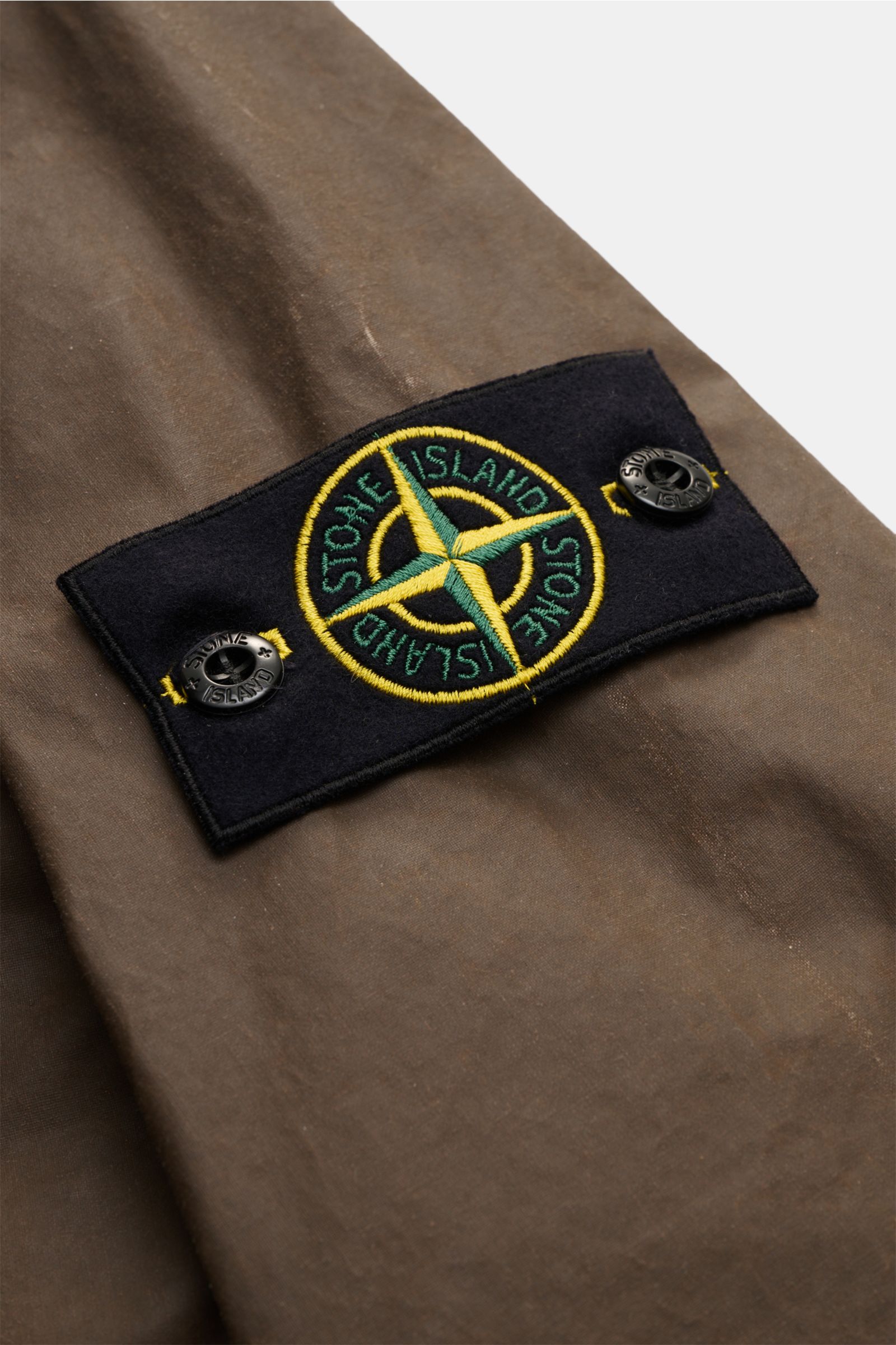 Stone Island Wachsjacke 'Waxed Pigment Cotton Tela' khaki im Detail, Nahaufnahme des typischen Kompass-Badges auf gewachster, wasserabweisender Baumwoll-Polyester-Oberfläche.