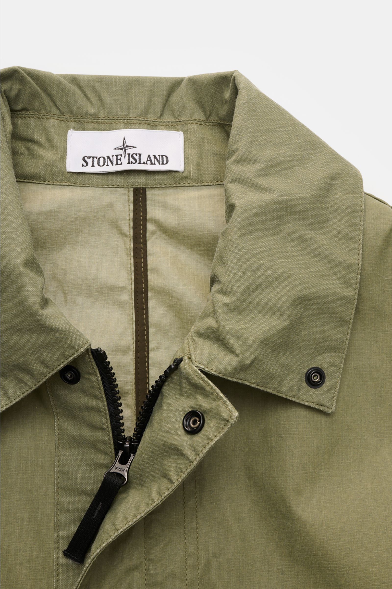 Stone Island Wachsjacke 'Waxed Pigment Cotton Tela' oliv shown from a close-up front perspective, featuring a durable olive cotton-polyester canvas with a waxed, water-repellent surface and robust YKK zipper. 

Innovativ wie gewohnt und prädestiniert f