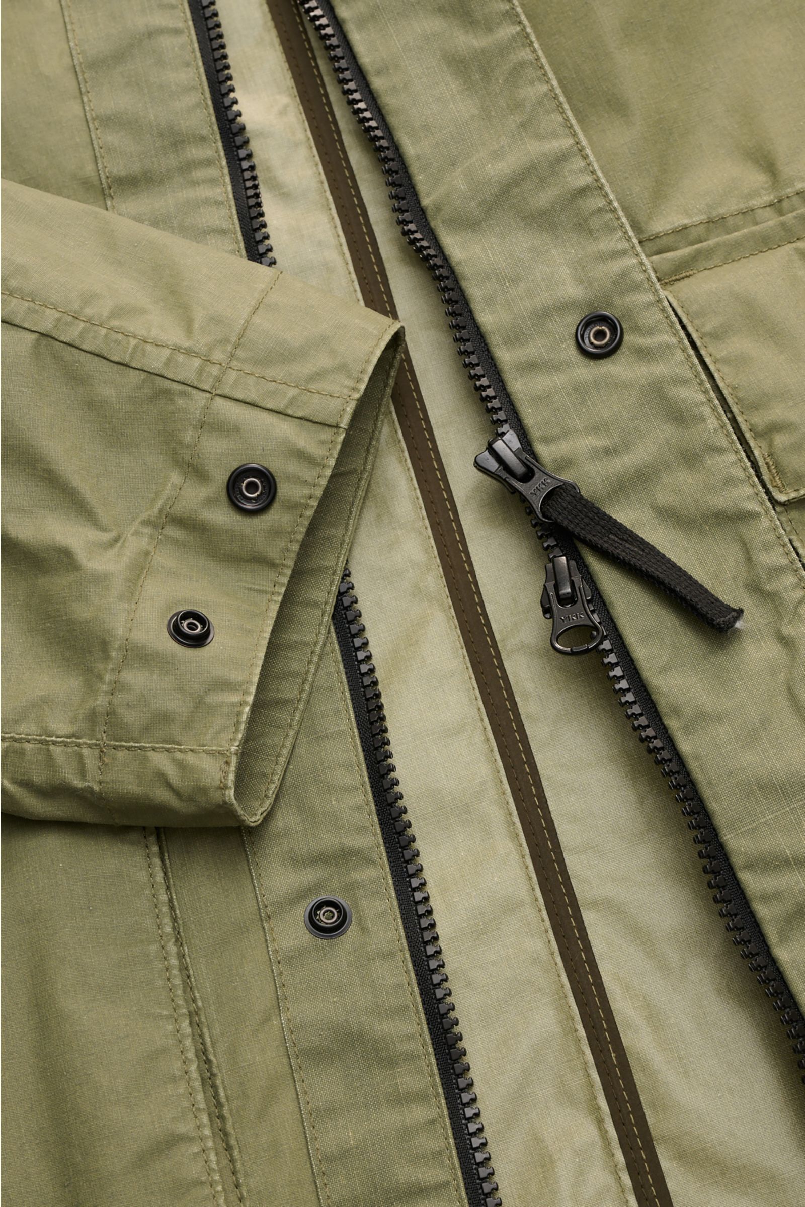 Close-up front view of Stone Island Wachsjacke 'Waxed Pigment Cotton Tela' oliv showing zipper, snap buttons, and olive green fabric texture.

Description: Innovativ wie gewohnt und prädestiniert für unbeständiges Wetter ist die Wachsjacke 'Waxed Pigme