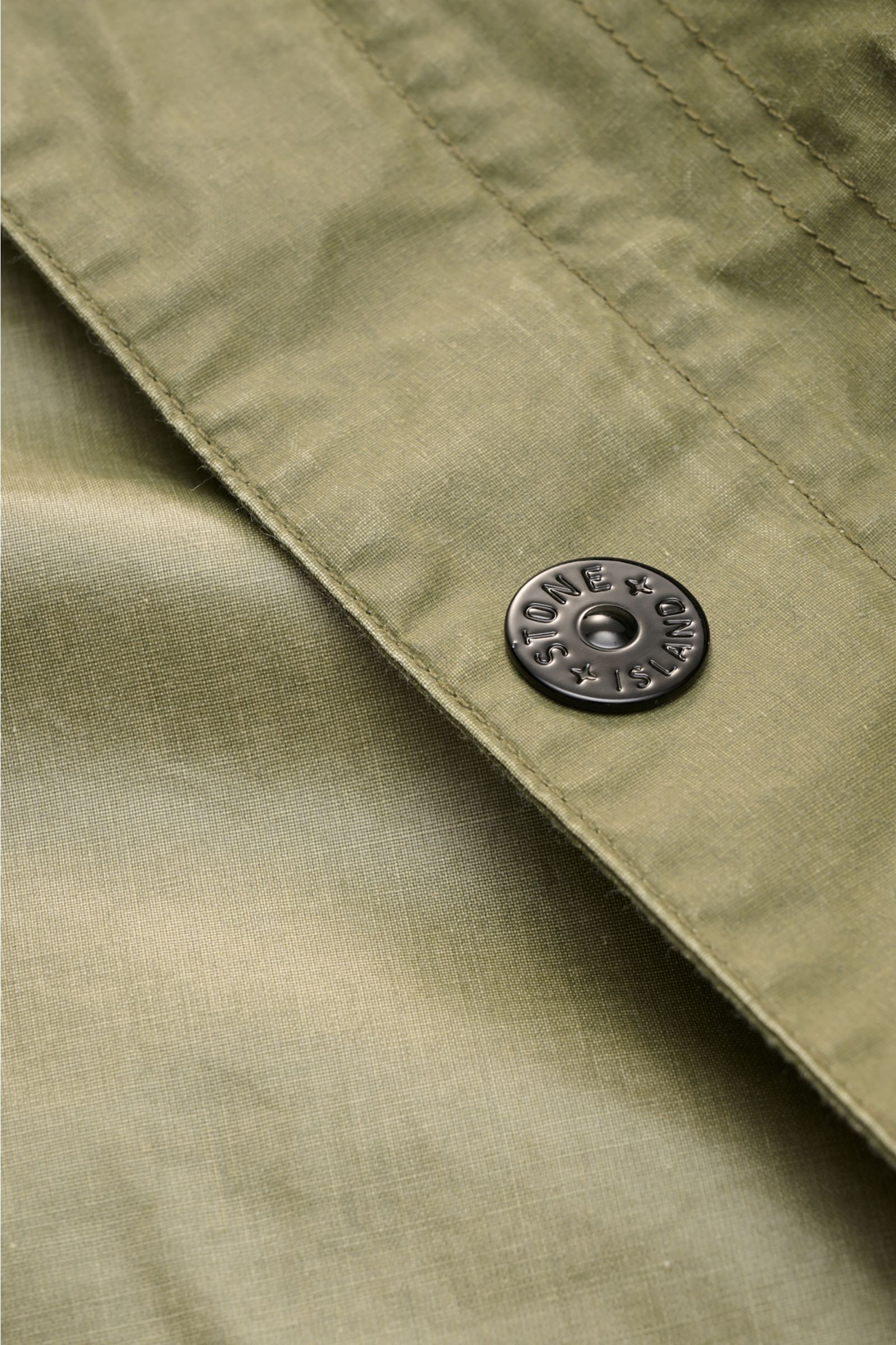 Close-up, angled view of the olive Stone Island Wachsjacke 'Waxed Pigment Cotton Tela' showing the waxed cotton-polyester fabric and a black snap button engraved with 'STONE ISLAND'; innovativ wie gewohnt und prädestiniert für unbeständiges Wetter ist die