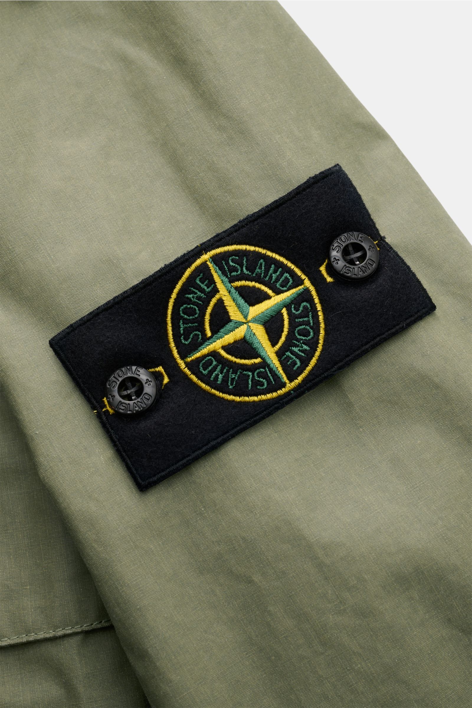 Stone Island Wachsjacke 'Waxed Pigment Cotton Tela' oliv, Nahaufnahme des Ärmel-Logos aus gewachster Baumwolle mit robustem Griff, wasserabweisend.