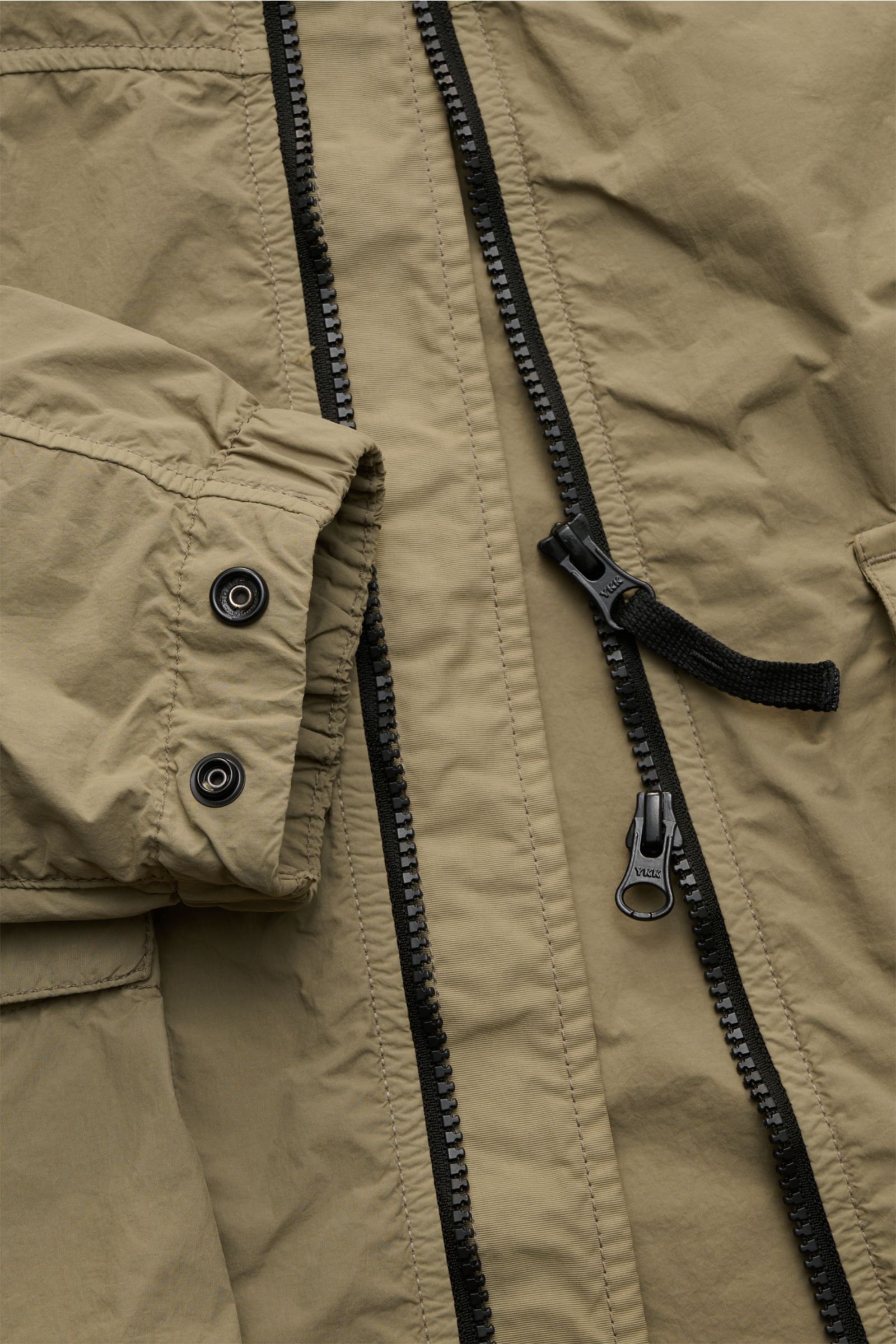 Nahaufnahme der Stone Island Jacke 'Nylon Smerigliato-TC' oliv, von oben auf Reißverschluss, Ärmel mit Druckknöpfen und Materialstruktur.