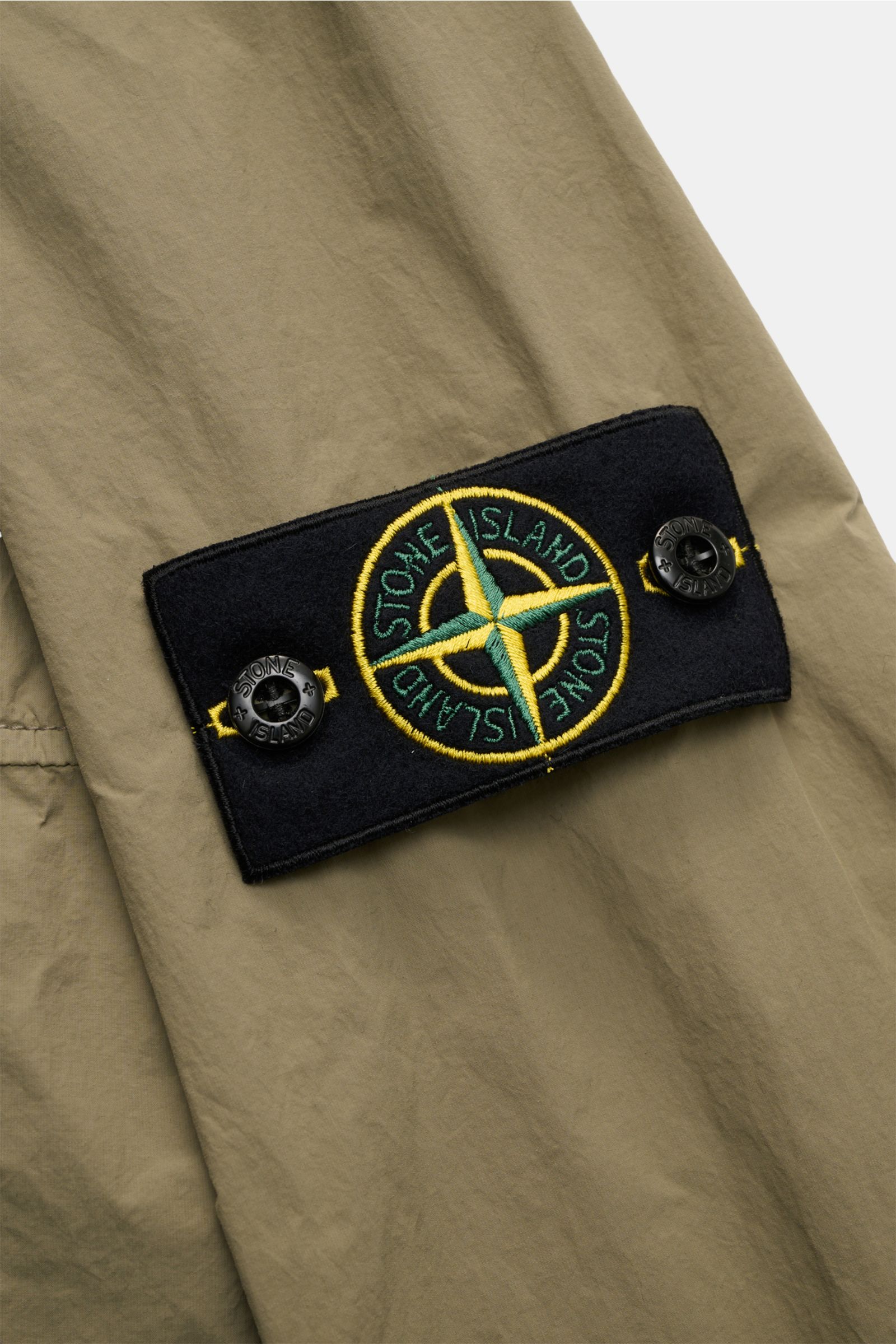 Nahaufnahme des Ärmelbereichs der Stone Island Jacke 'Nylon Smerigliato-TC' oliv mit schwarzem Kompass-Badge, Knöpfen und matter Oberfläche.