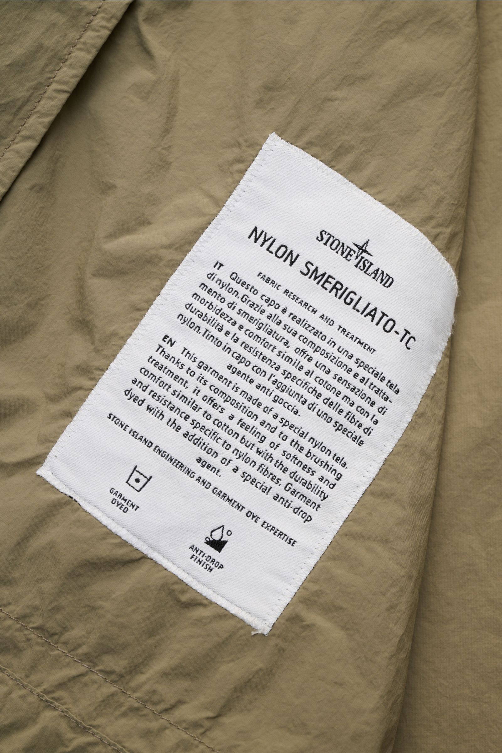 Nahaufnahme des weißen Labels der Stone Island Jacke 'Nylon Smerigliato-TC' oliv mit Material- und Pflegehinweisen auf olivgrünem Nylonstoff.