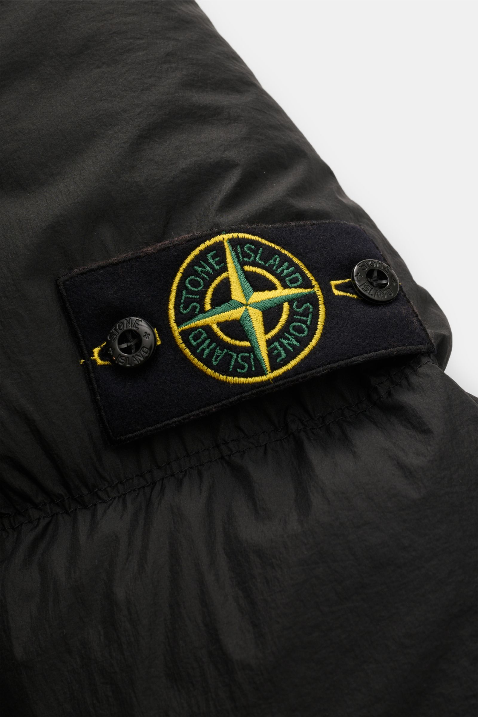 Close-up of the sleeve with logo patch of the Stone Island Daunenjacke 'Seam Faded Light Nylon Tela' schwarz, photographed from the side showing the black microfiber fabric and embroidered emblem.

Stilsicher auch an kalten Tagen: Die Daunenjacke von S