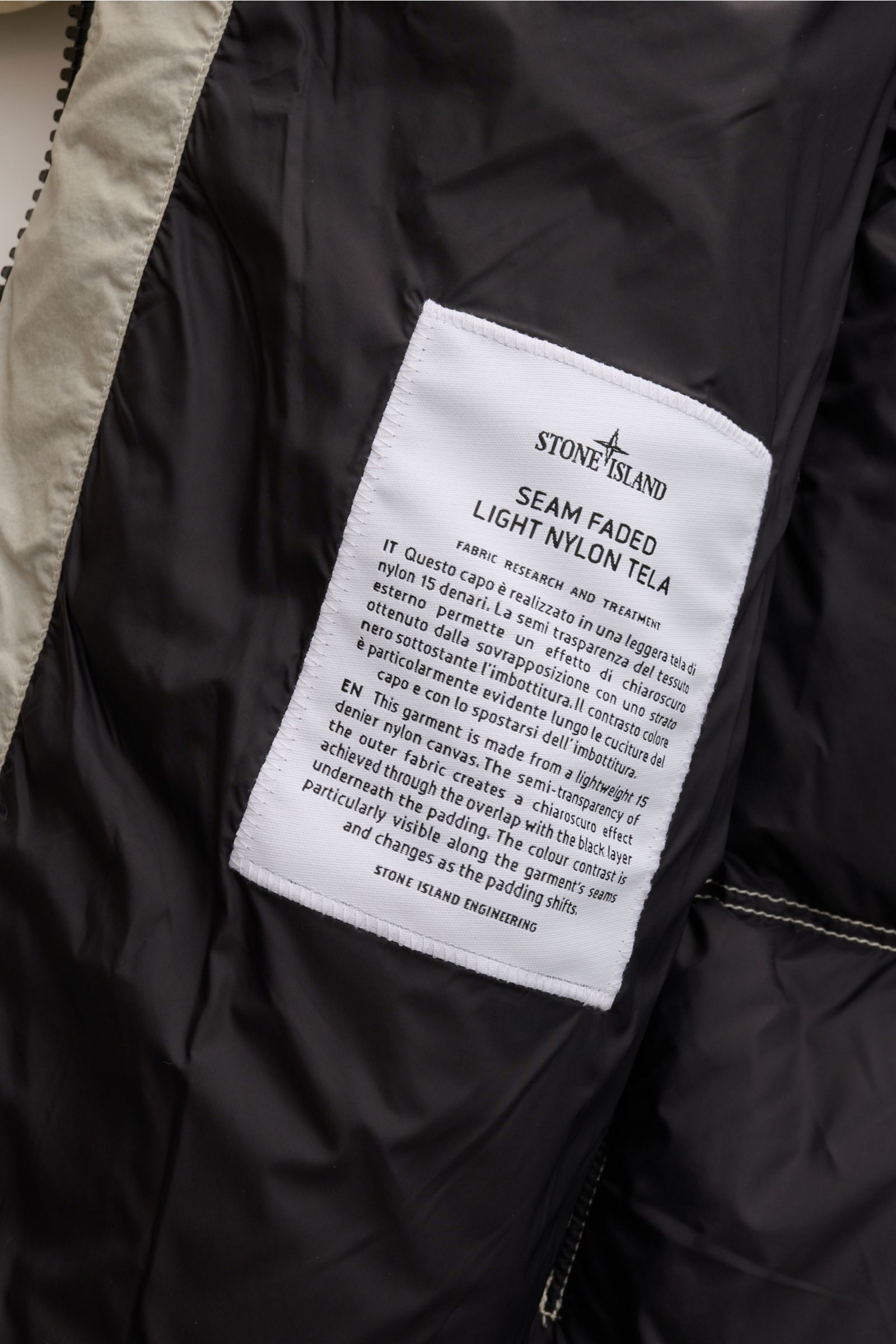 Innenansicht der Stone Island Daunenjacke 'Seam Faded Light Nylon Tela' offwhite mit schwarzem Futter, Nahtdetail und weißem Markenlabel.
