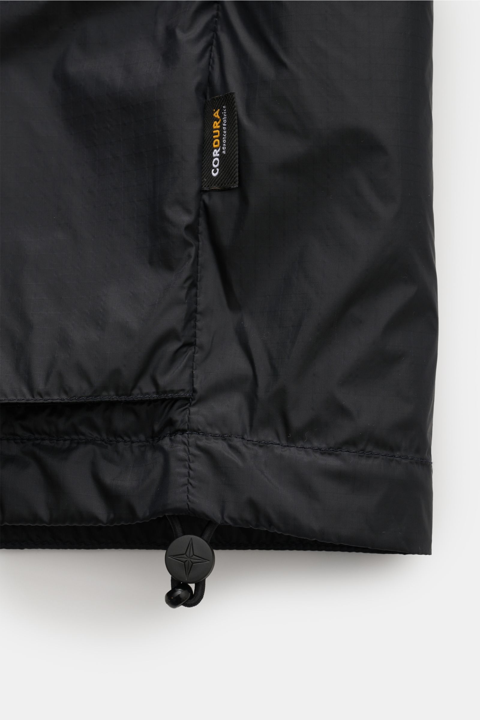 Close-up lower view of the Stone Island Blouson 'Light Ripstop Cordura' black, showing high-strength ripstop fabric, elastic hem with drawstring and branded toggle, and Cordura label. Robust yet lightweight, made from high-quality microfibre with a smooth