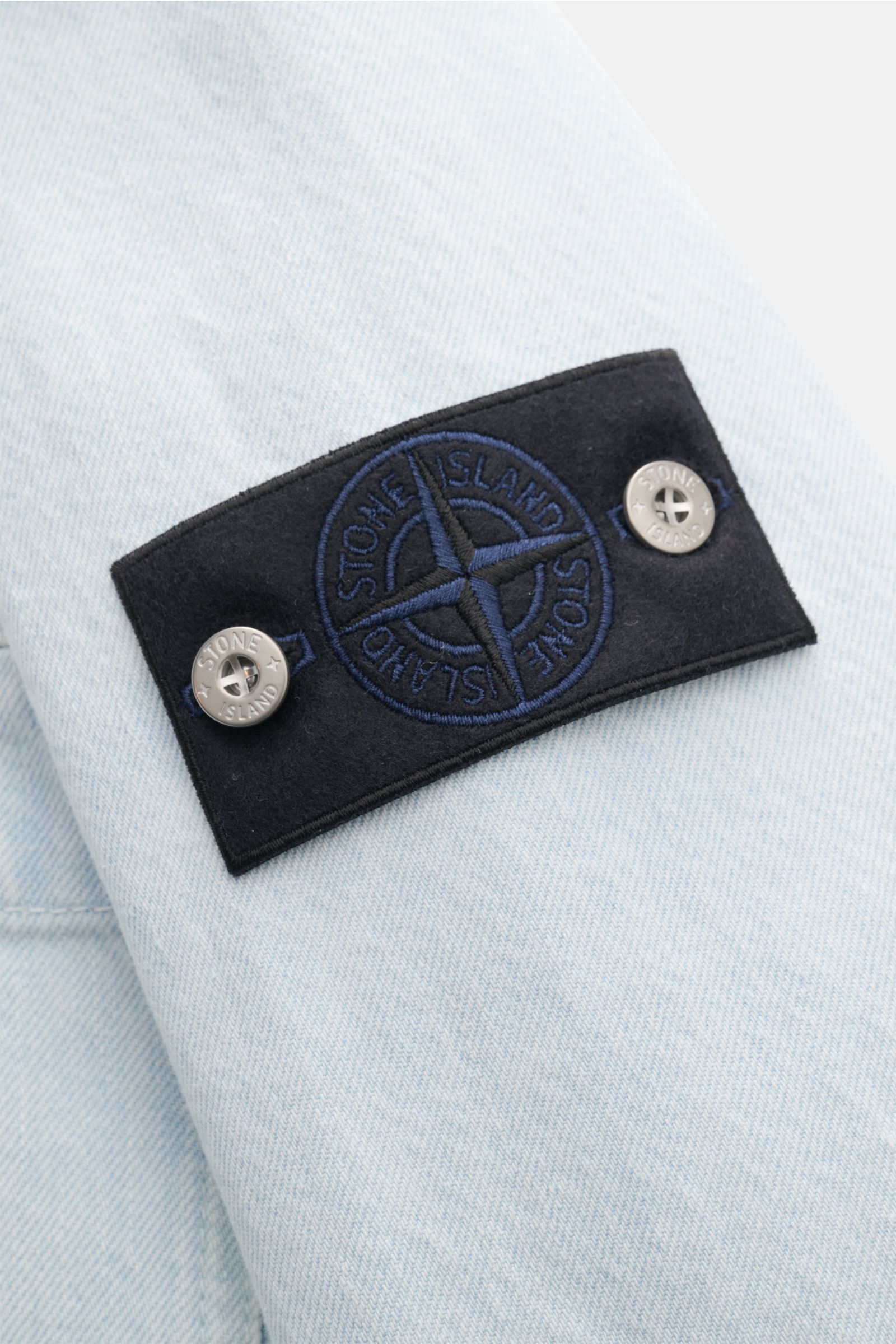 Close-up angled view of the Stone Island Denim jacket pastel blue sleeve featuring the black compass badge with blue embroidery and two silver buttons, highlighting the denim’s typical fabric texture and craftsmanship. A timeless companion for urban sprin