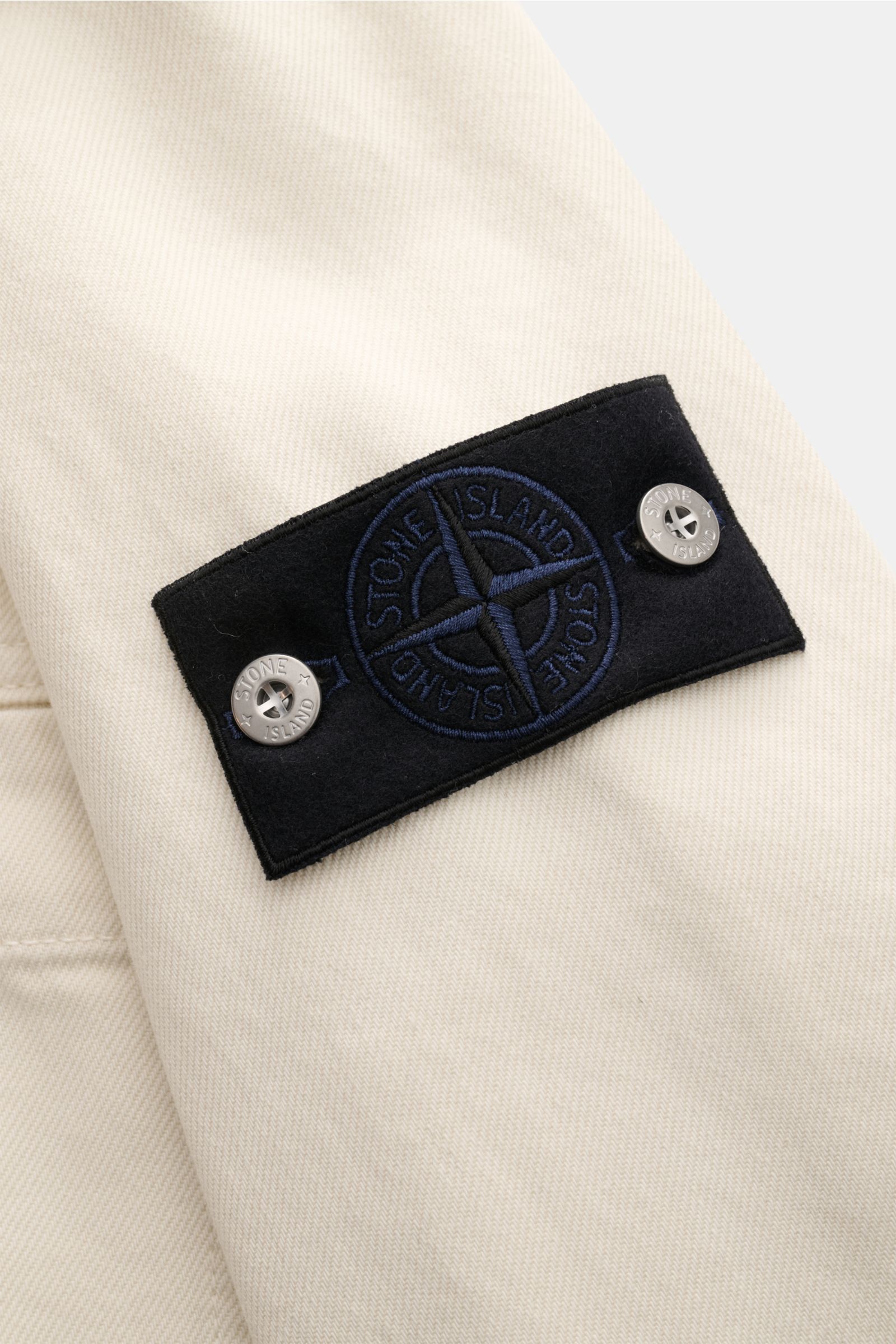 Stone Island Jeansjacke 'Light Bull Denim-TC' creme, Detailansicht des schwarzen Kompass-Badges auf leichtem, wasserabweisendem Denim.