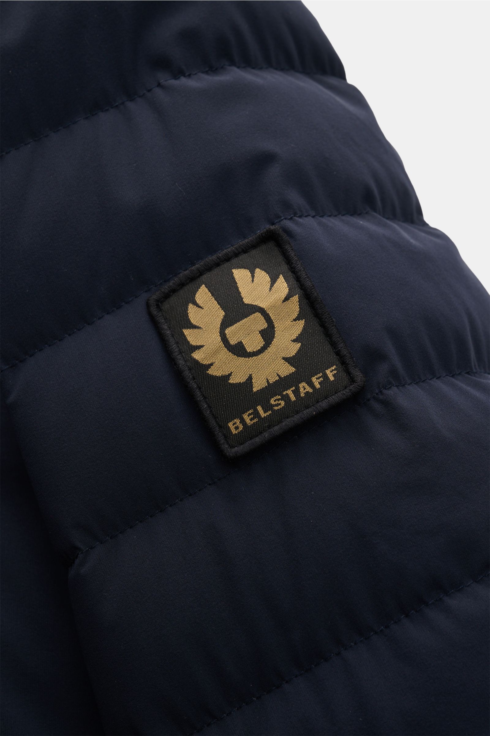 Nahaufnahme der Belstaff Daunenjacke 'Chassis' navy von schräg oben, zeigt das BELSTAFF-Weblabel auf hochwertiger, wasserabweisender Microfaser mit leichter Daunenfüllung.