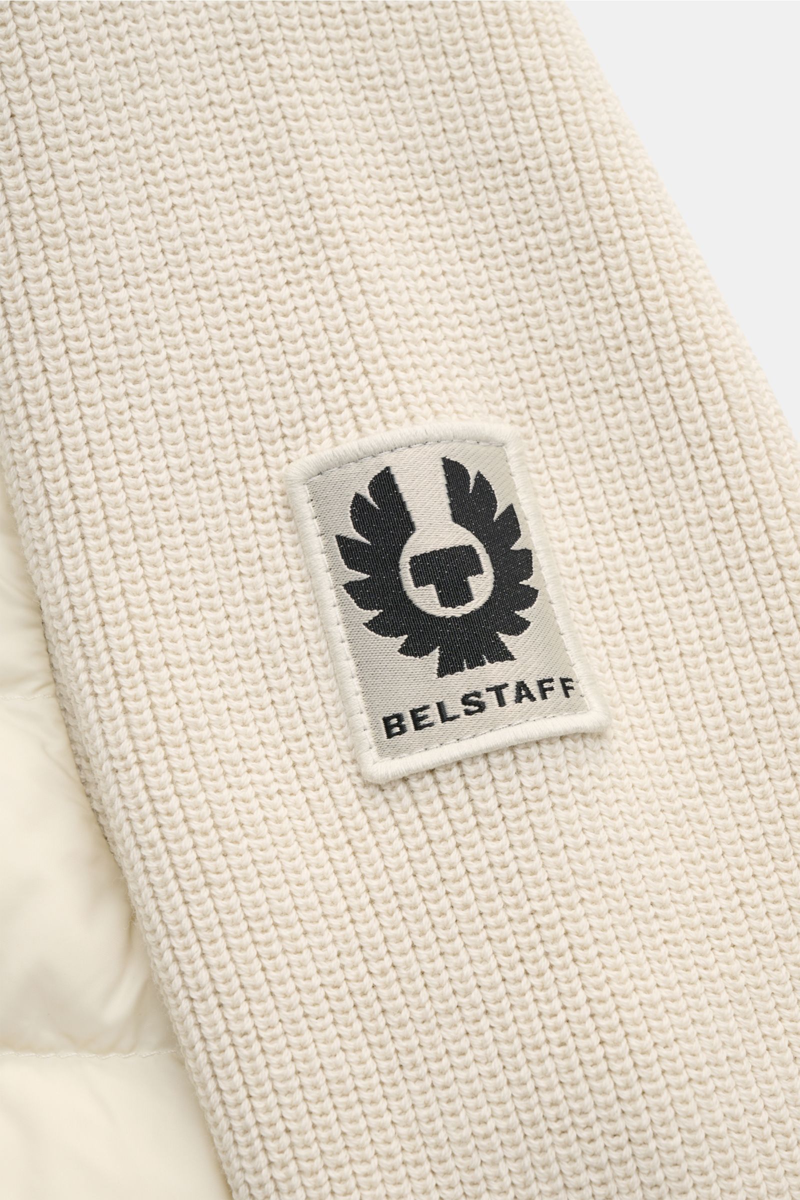 Nahaufnahme der Belstaff Daunenjacke 'Control Cardigan' sand, strickiger Wollärmel mit BELSTAFF-Weblabel am Oberarm, gesteppte Daunenfront.