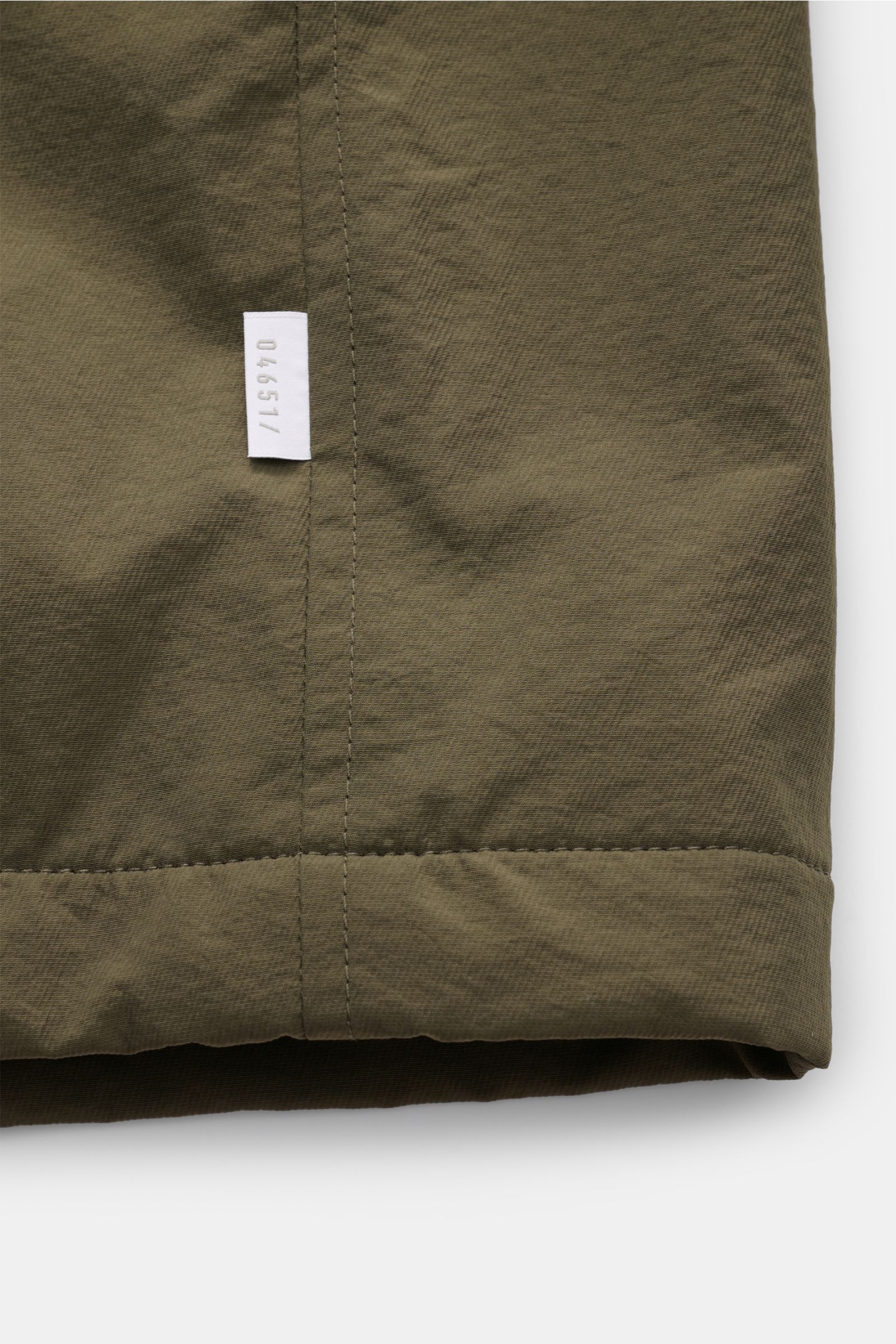 Close-up view of the hem and side seam of the 04651/ A trip in a bag Jacket olive, showing high-quality microfibre fabric with a small white woven label. The jacket features a smooth feel, lightweight padding, wind and water repellent, regular fit, zip, c