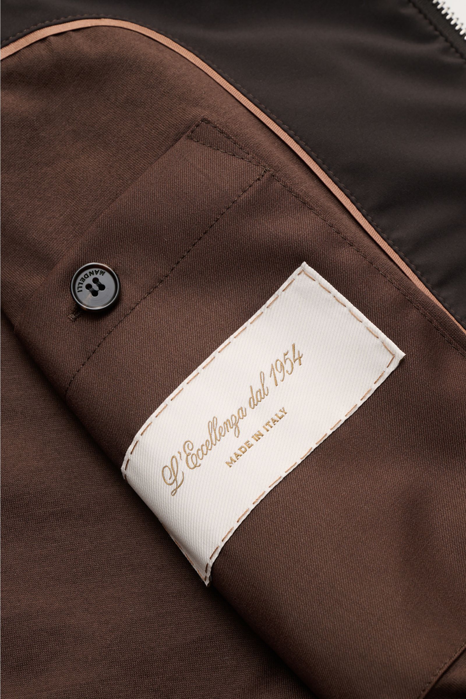 Close-up interior view of the Mandelli Blouson dark brown showing a brown fabric pocket with a black Mandelli button and a white label reading 'L’Eccellenza dal 1954 Made in Italy.' A perfect symbiosis of luxury and comfort: This blouson by MANDELLI is co