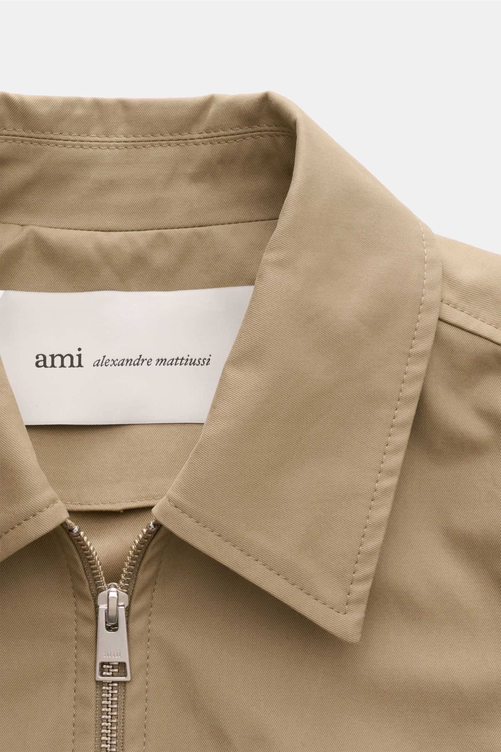 Close-up front view of the Ami Paris Blouson taupe showing the collar, two-way zipper with 'ami' engraving, and the label inside the collar. Stilvoller Begleiter in der Übergangszeit: Der AMI PARIS Blouson vereint legeren Stil mit hohem Tragekomfort und b