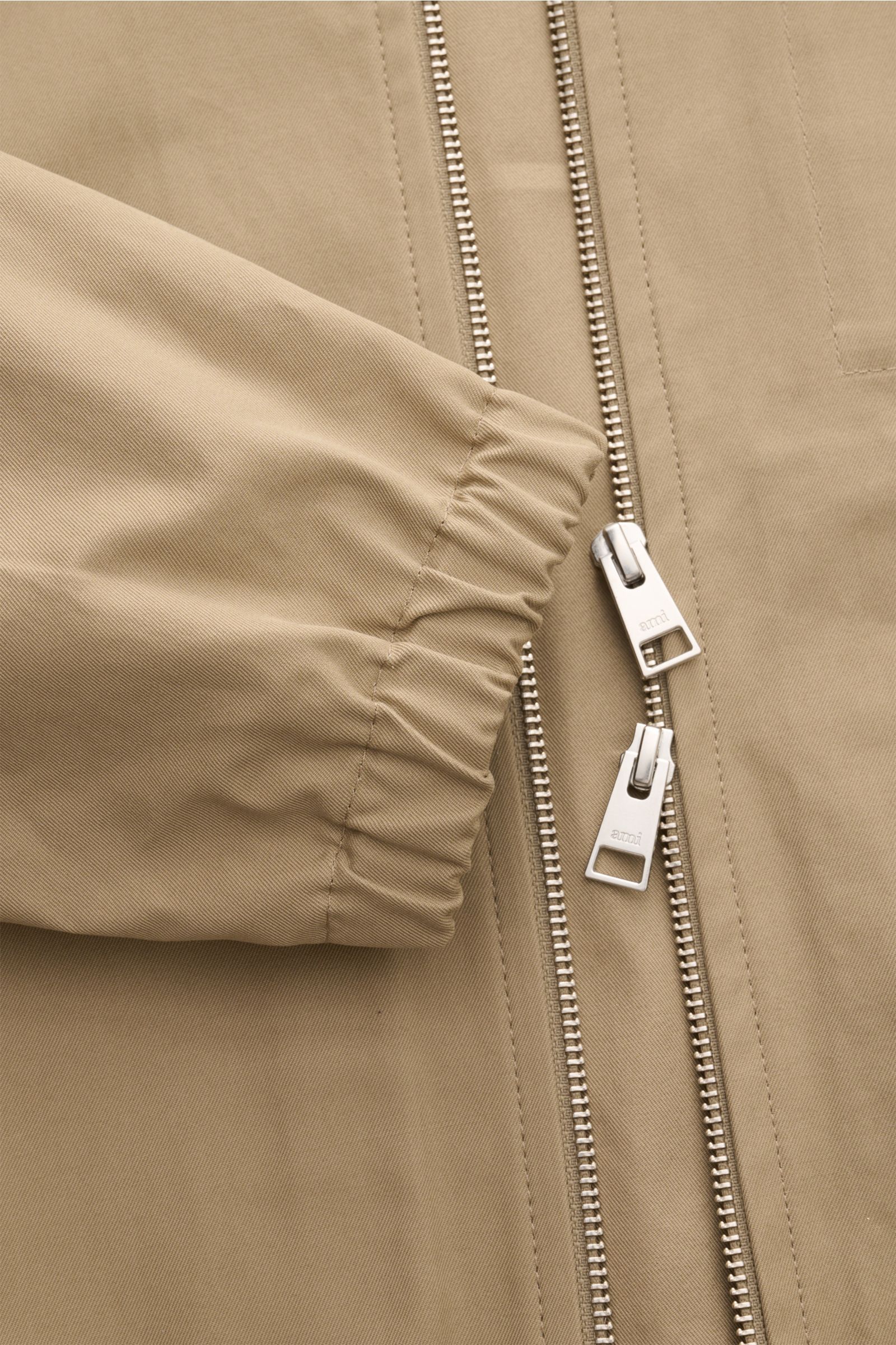 Close-up front view of the Ami Paris Blouson taupe showing its two-way zipper with logo, elastic sleeve cuff, and smooth cotton fabric.
