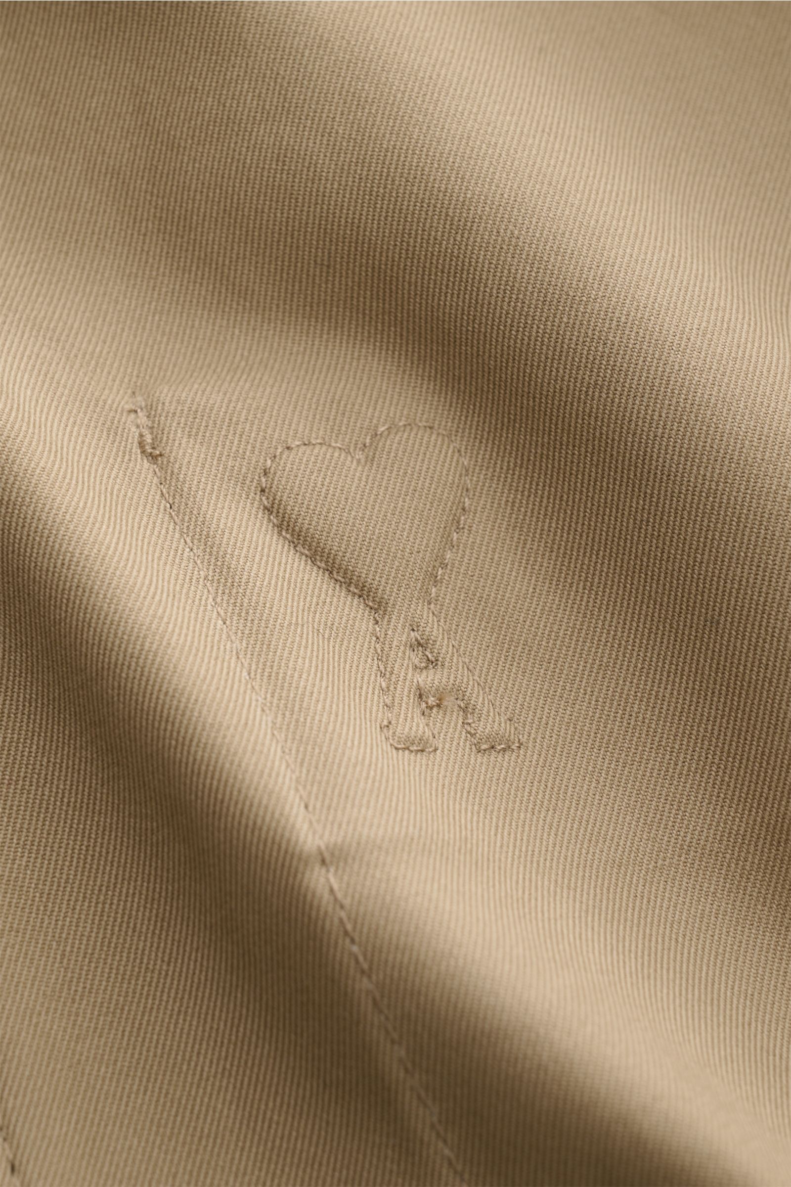 Close-up front detail of taupe Ami Paris Blouson showing cotton fabric, logo stitching, and texture from a slightly angled perspective. Stilvoller Begleiter in der Übergangszeit: Der AMI PARIS Blouson vereint legeren Stil mit hohem Tragekomfort und beglei