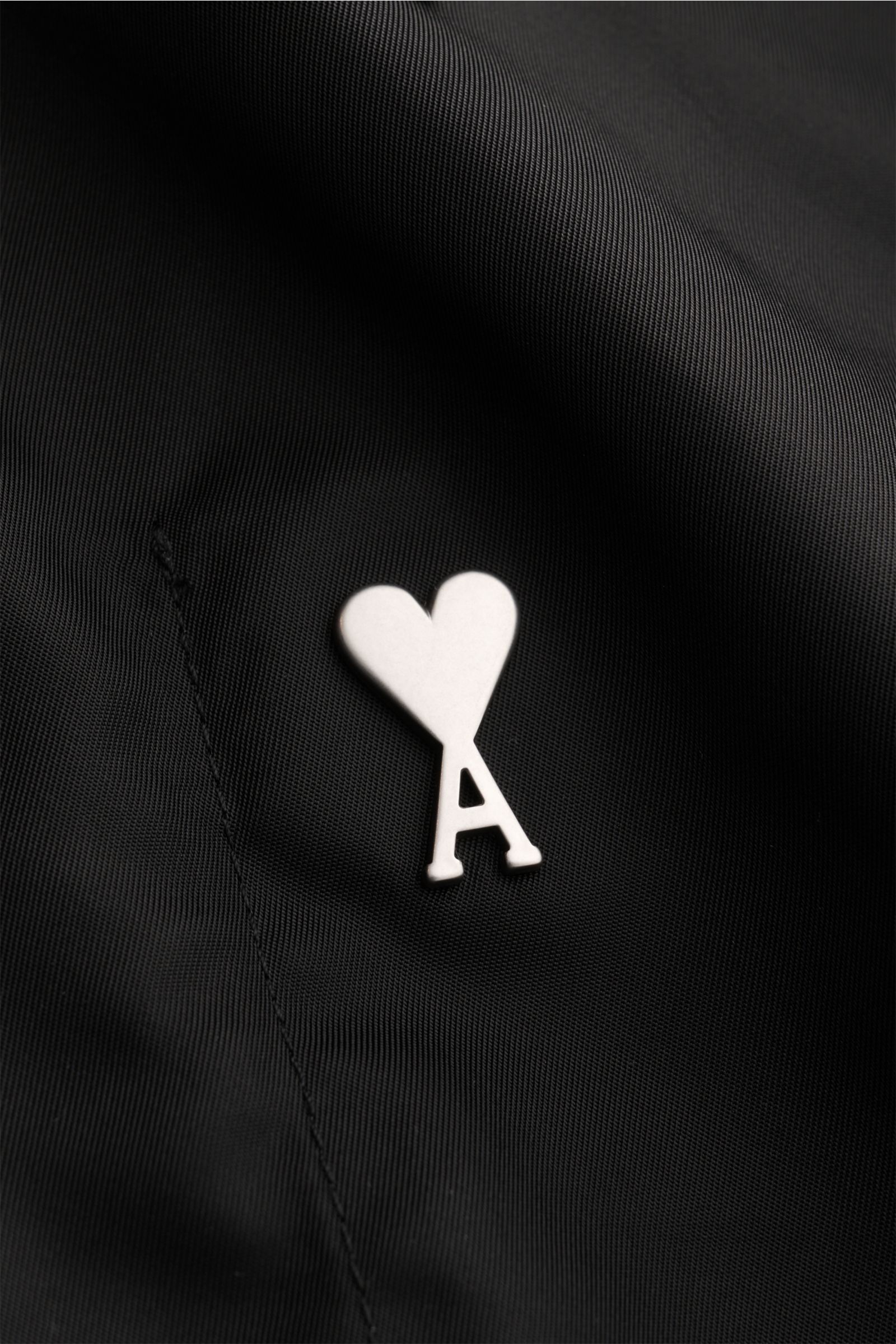 Close-up front view of the Ami Paris Jacke schwarz showing the brand's heart-shaped 'A' logo on smooth, high-quality black microfiber fabric. Stylish transitional jacket with regular fit, snap button placket, turn-down collar, piped front pockets with sna