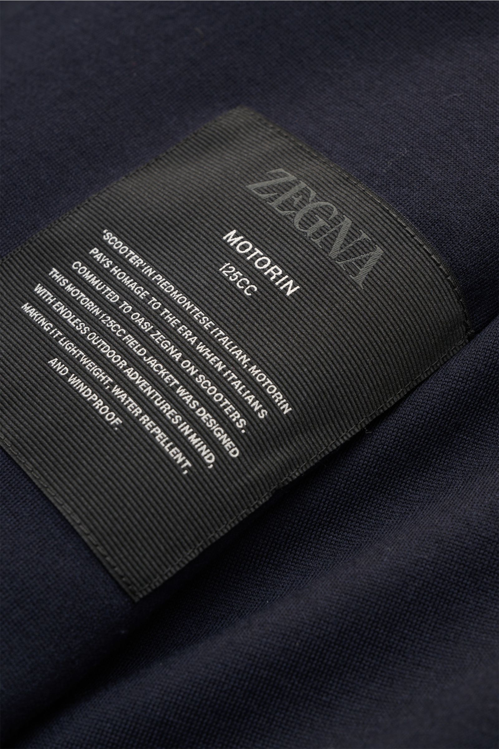 Close-up of the Zegna Jacket 'Motorin' navy label on the soft, high-quality microfibre fabric, photographed from above showing detailed text. 