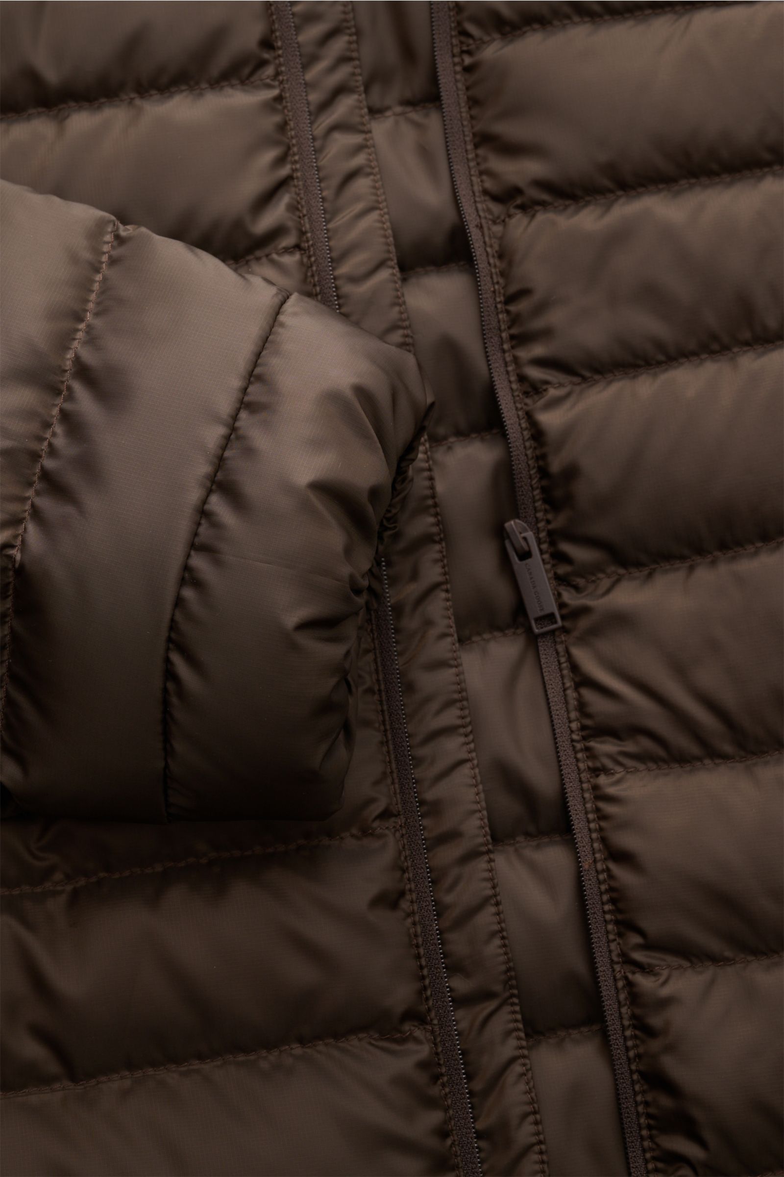 Nahaufnahme der dunkelbraunen Canada Goose Daunenjacke 'Stratus Tonal' mit Ripstop-Obermaterial, Stehkragen, Zwei-Wege-Reißverschluss und elastischem Ärmelbündchen, von schräg oben fotografiert.