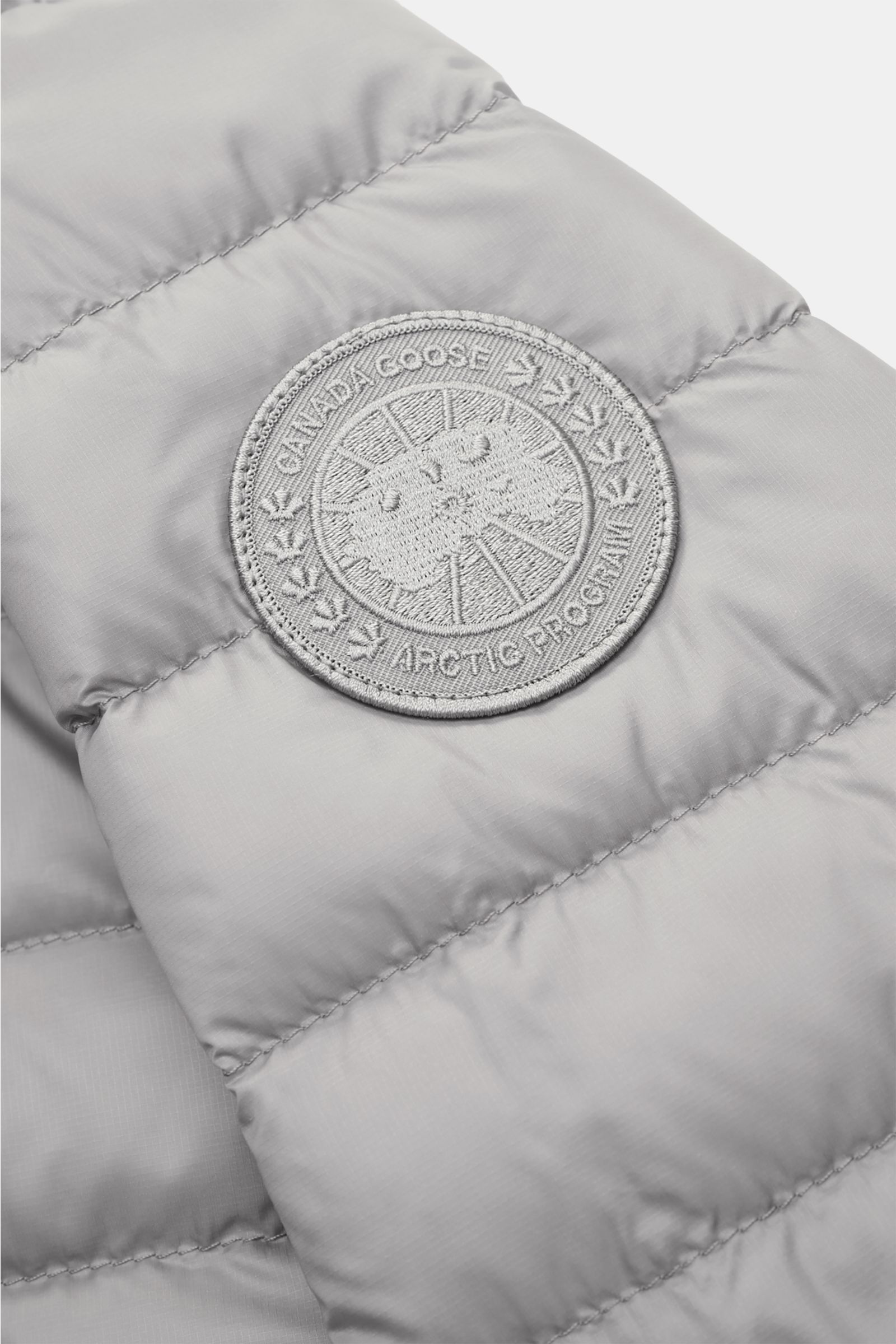 Nahaufnahme der Canada Goose Daunenjacke 'Stratus Tonal' hellgrau von schräg oben, zeigt Ripstop-Obermaterial und tonales Weblabel am Oberarm.