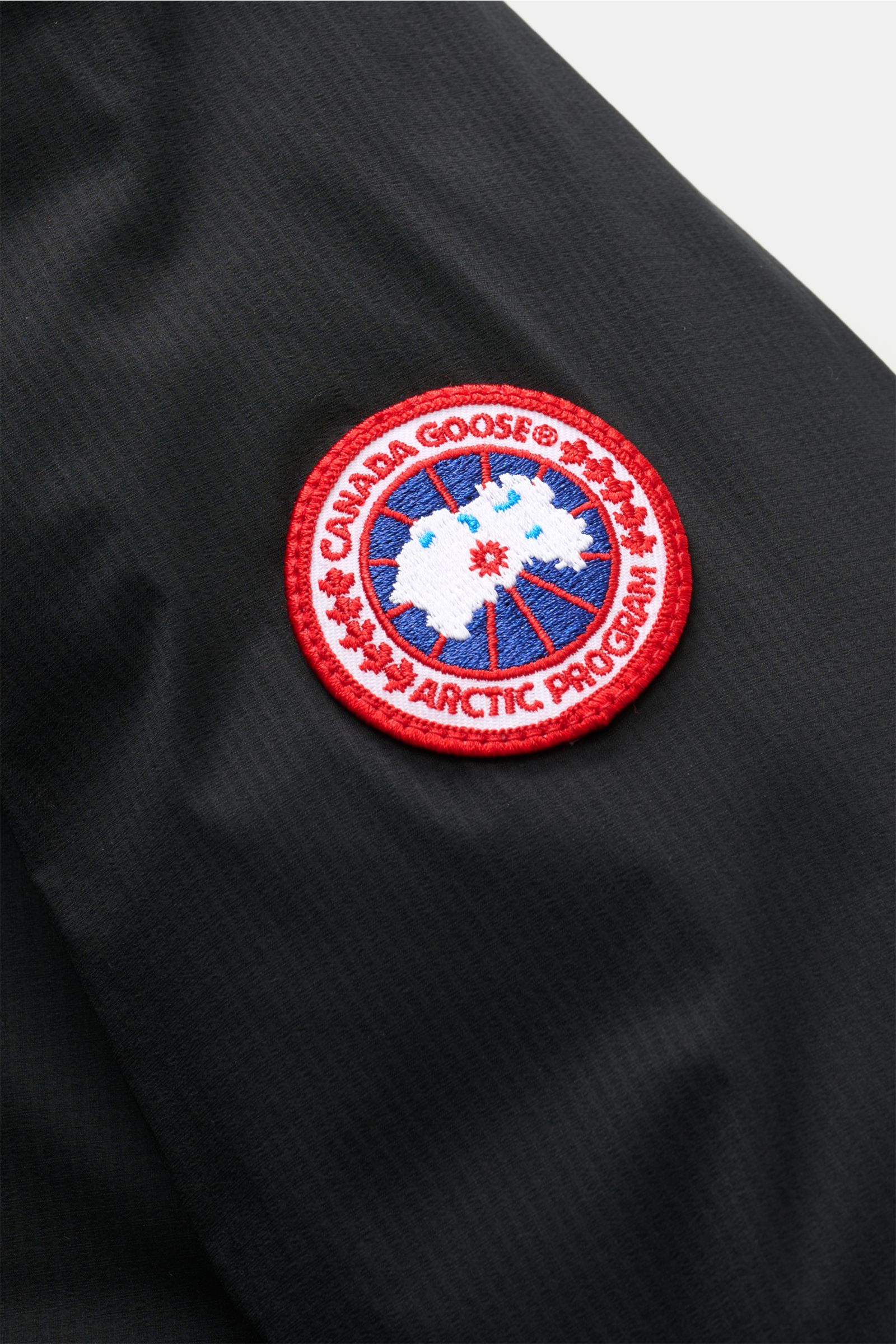 Close-up side view of Canada Goose Daunenjacke 'Lodge Hoody' schwarz showing the red, white, and blue Arctic Program patch on black ripstop microfiber fabric.

Maximaler Komfort in leichter Ausführung: Die in sich verstaubare Daunenjacke 'Lodge Hoody' 