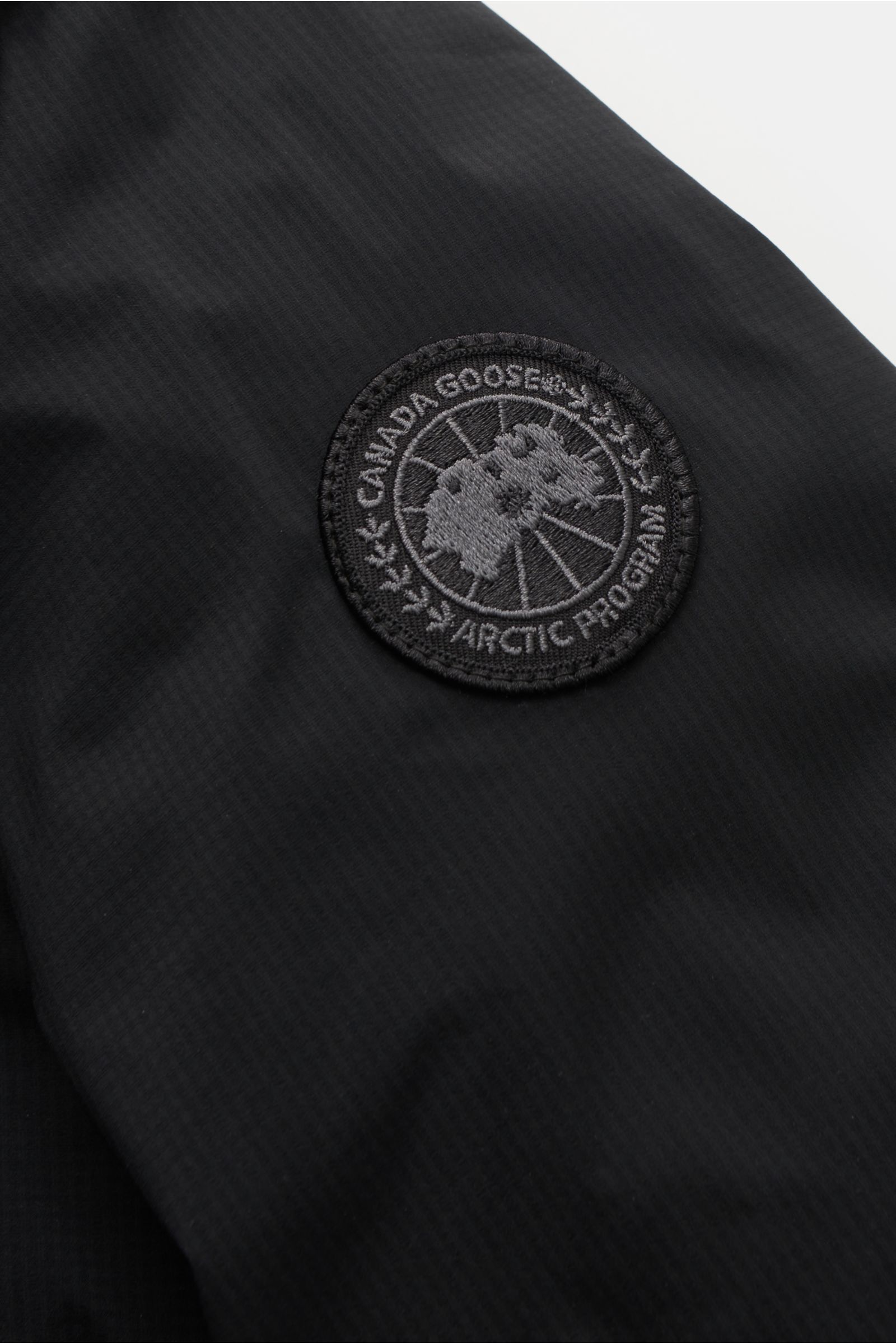 Close-up of the tonal Canada Goose Daunenjacke 'Lodge Hoody' schwarz logo patch on black ripstop fabric, photographed from above.

Description: Maximaler Komfort in leichter Ausführung: Die in sich verstaubare Daunenjacke 'Lodge Hoody' von CANADA GOOSE