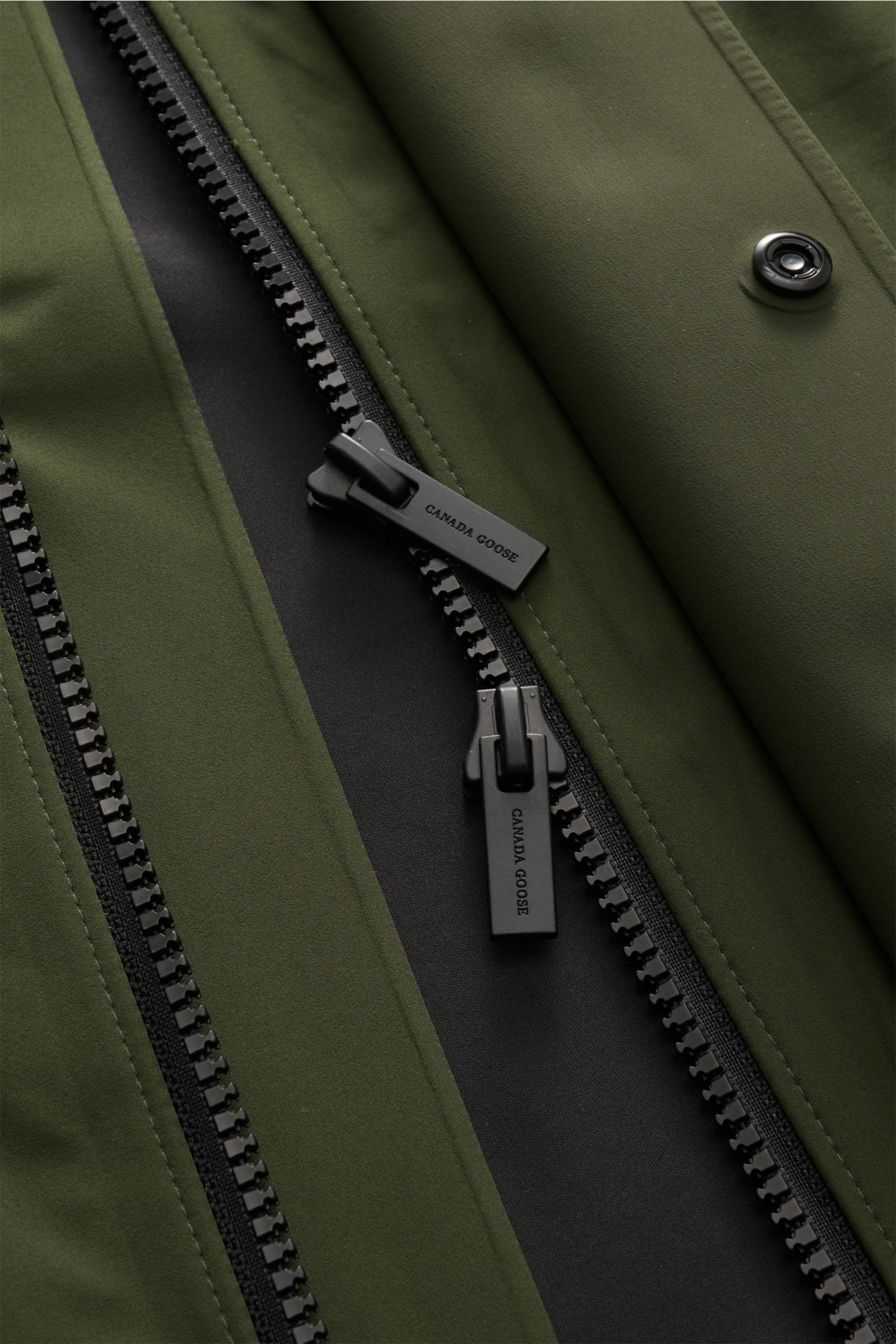 Nahaufnahme der Canada Goose Jacke 'Rupert' dunkelgrün, Fokus auf zwei-Wege-Reißverschluss und Druckknopf, glatte Microfaser.