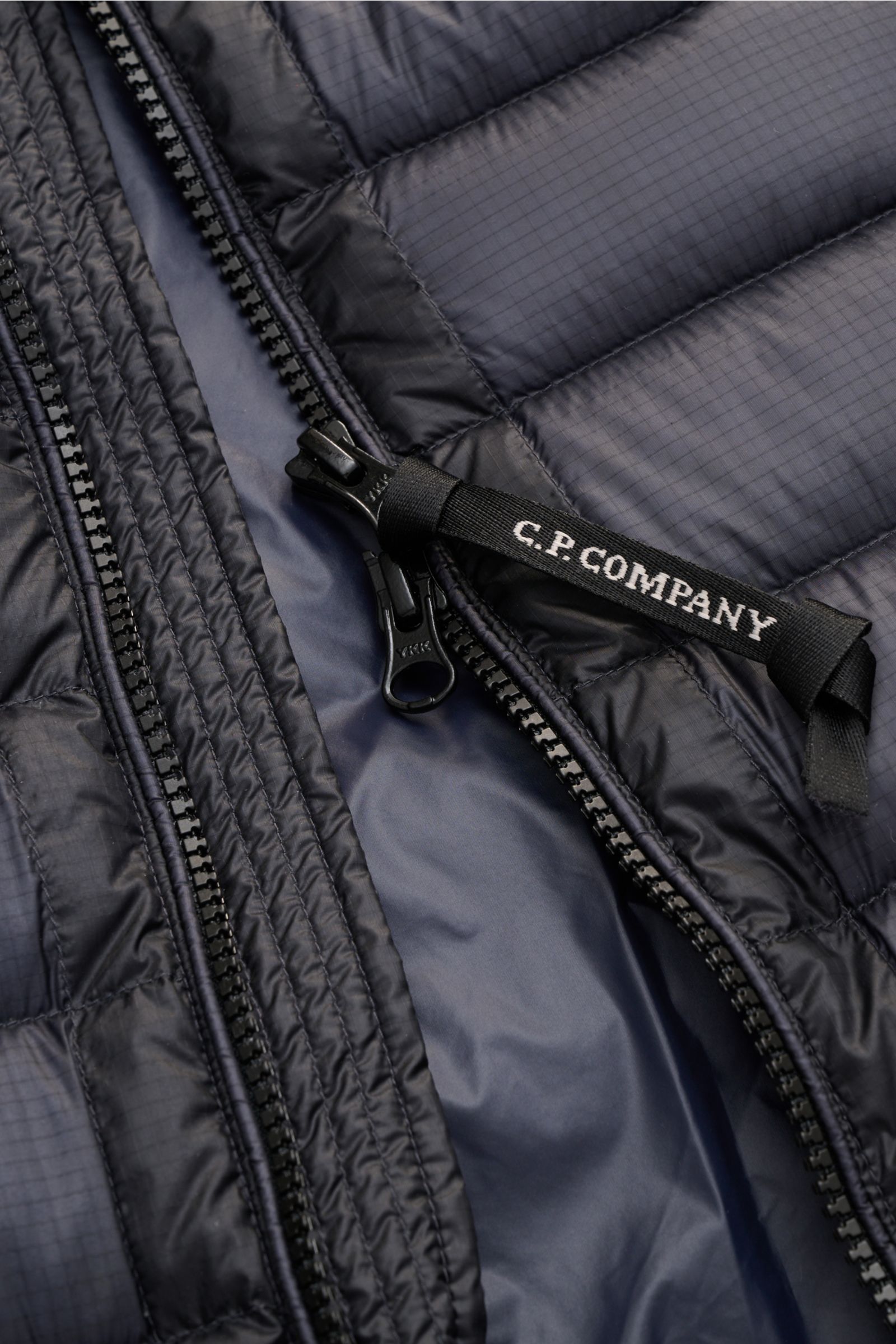 Nahaufnahme des C.P. Company Daunenblouson dark navy von innen, zeigt wasserabweisende Ripstop-Oberfläche, ultraleichte Daunenwattierung, robusten Zwei-Wege-Reißverschluss mit C.P. Company Logoband, tonales Futter und feine Steppungen.