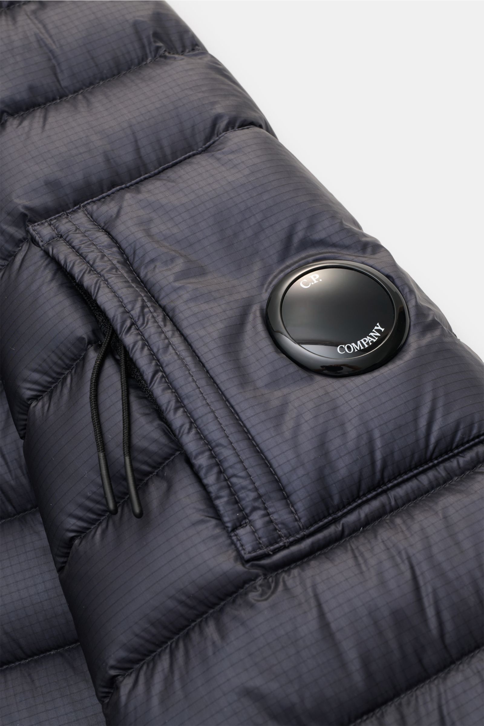 Nahaufnahme eines C.P. Company Daunenblouson dark navy von schräg oben, wasserabweisende Ripstop-Microfaser, ultraleichte Daunenwattierung, elastische Saum- und Ärmelabschlüsse, Kapuze mit Tunnelzug, robuste Reißverschlüsse, typisches Linsendetail am Ober