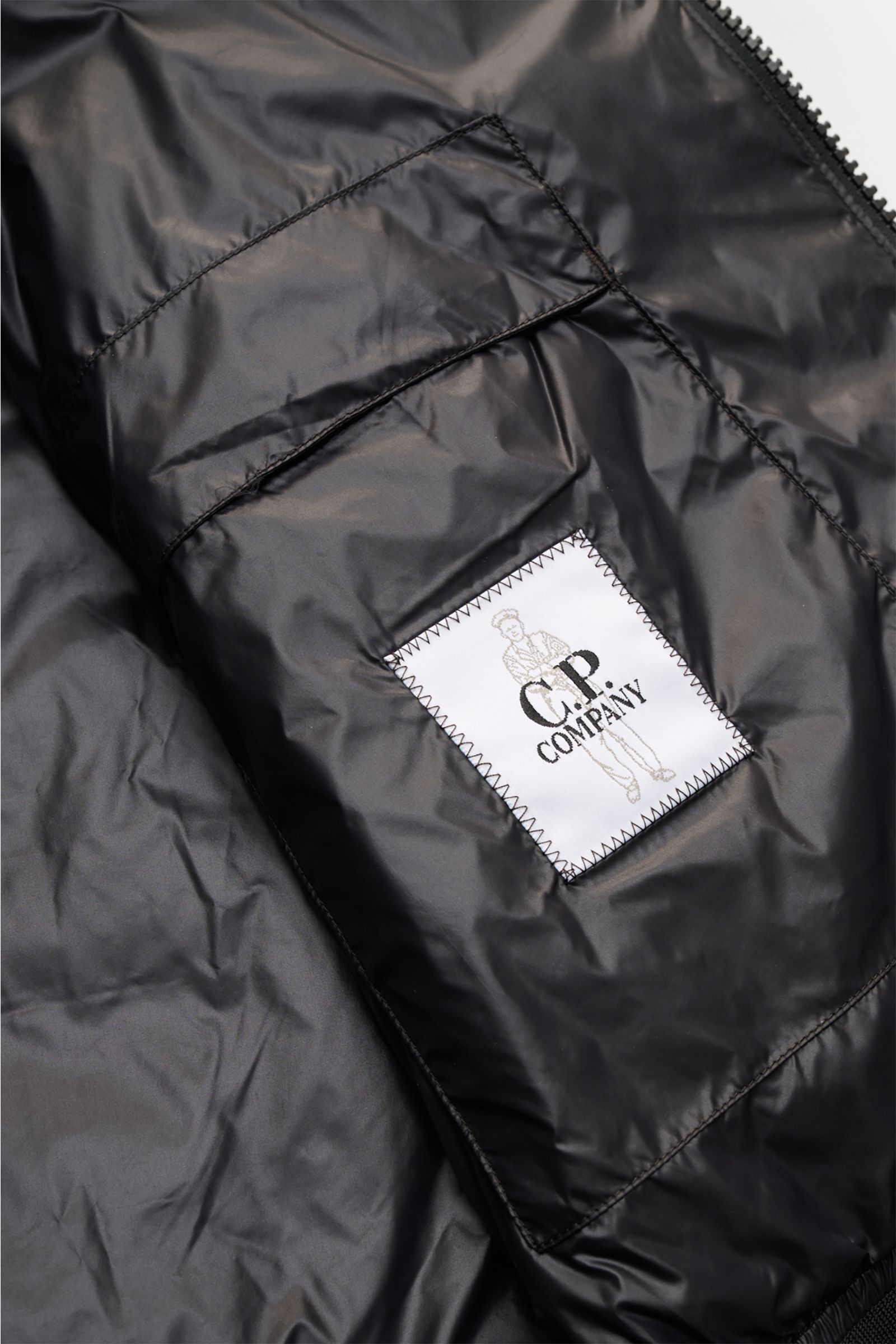 Close-up inside view of the black C.P. Company Down bomber jacket showing the stitched label on the inner pocket, highlighting technical lightness and water repellent ripstop surface.

Description: Technical lightness in its most modern form: This blouson