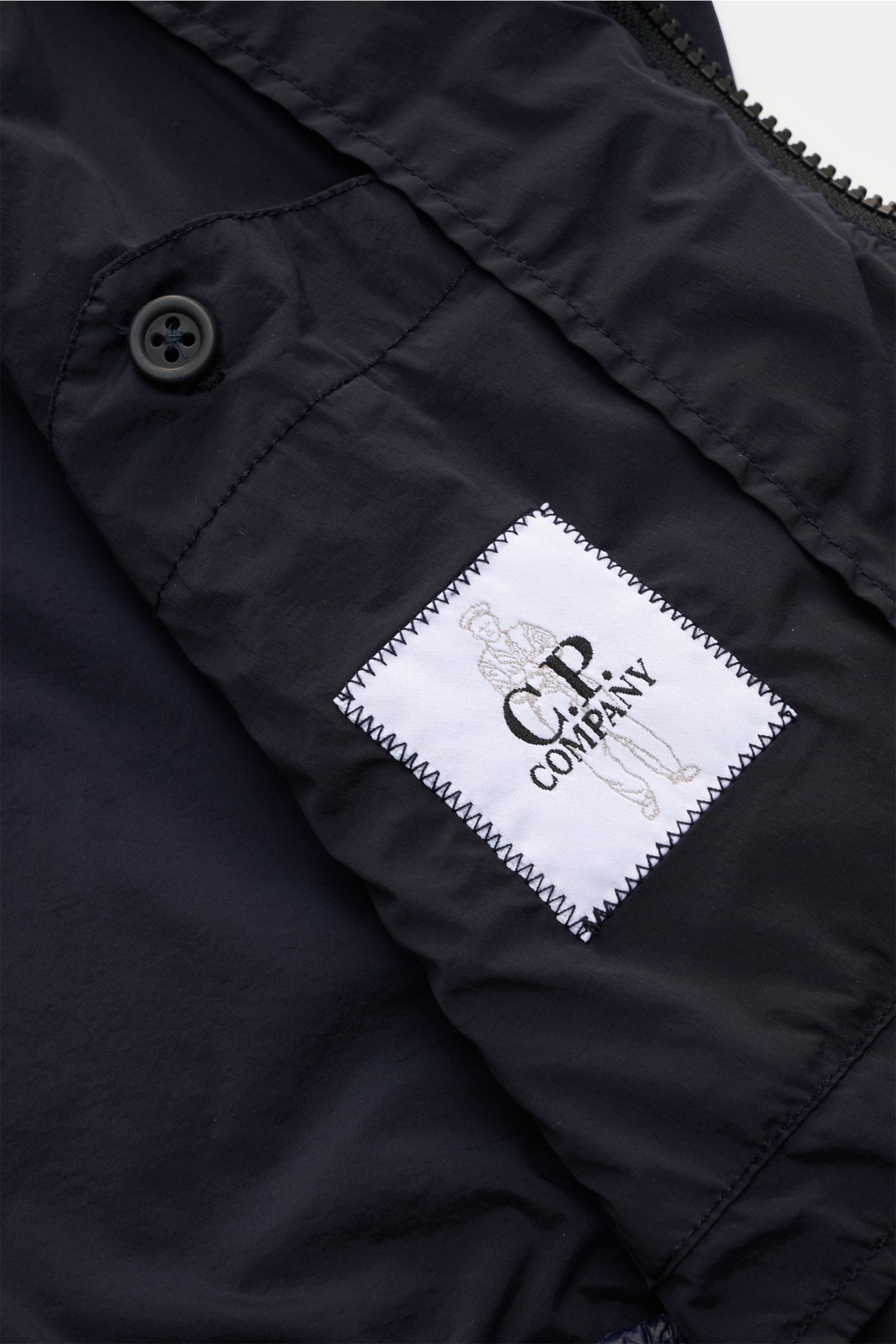 Nahaufnahme des Ärmels von C.P. Company Blouson 'Nycra R' dark navy aus recycelter Microfaser mit Label, Ärmeltasche und Knopf.