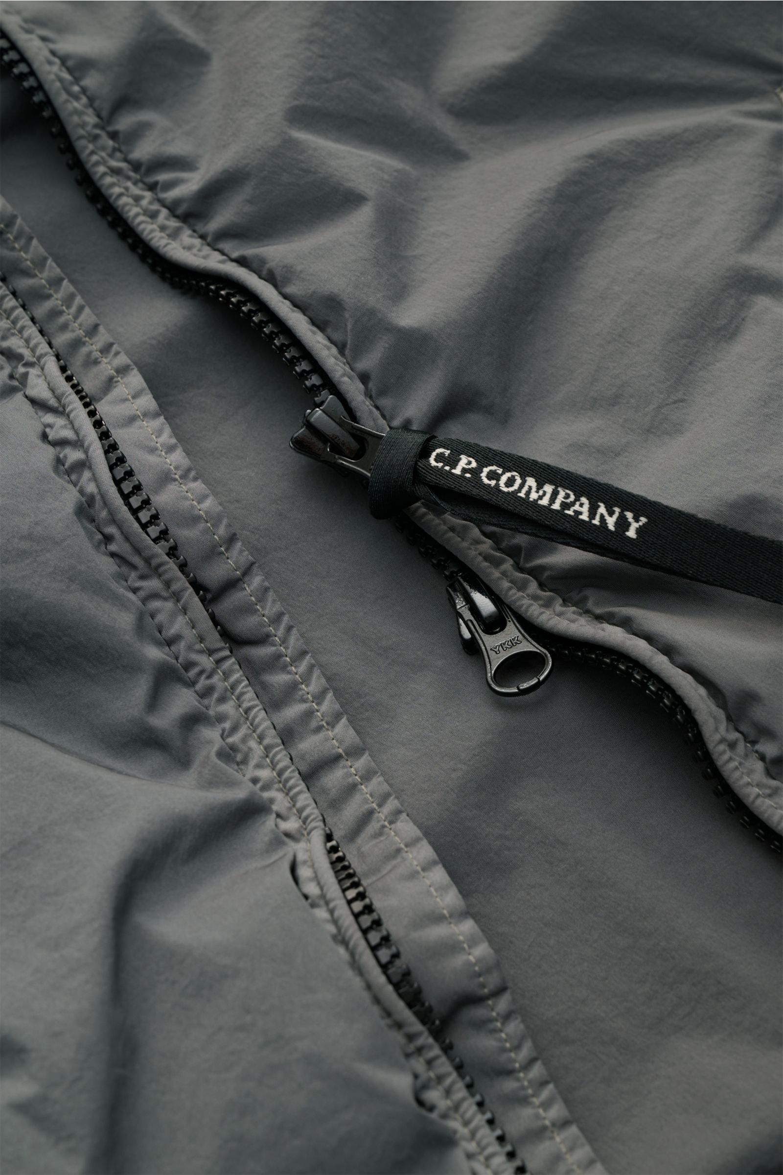 Nahaufnahme des grauen C.P. Company Blouson 'Nycra R' mit robustem Zwei-Wege-Reißverschluss und C.P. Company Logo-Schlaufe, glatte recycelte Microfaser, wind- und wasserabweisend, Slim Fit, komfortable Rippbündchen erkennbar.