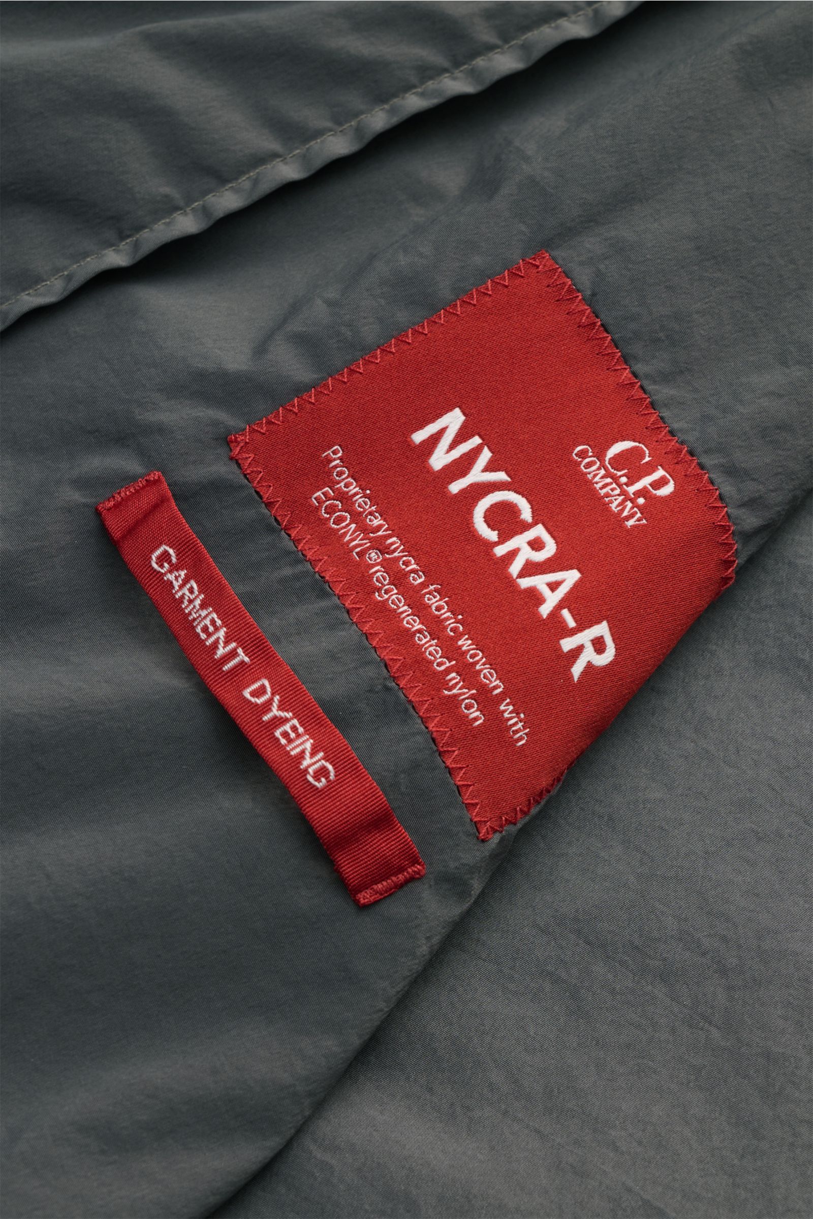 Nahaufnahme des roten Labels auf dem grauen C.P. Company Blouson 'Nycra R' grau, gefertigt aus recycelter, windabweisender Microfaser.