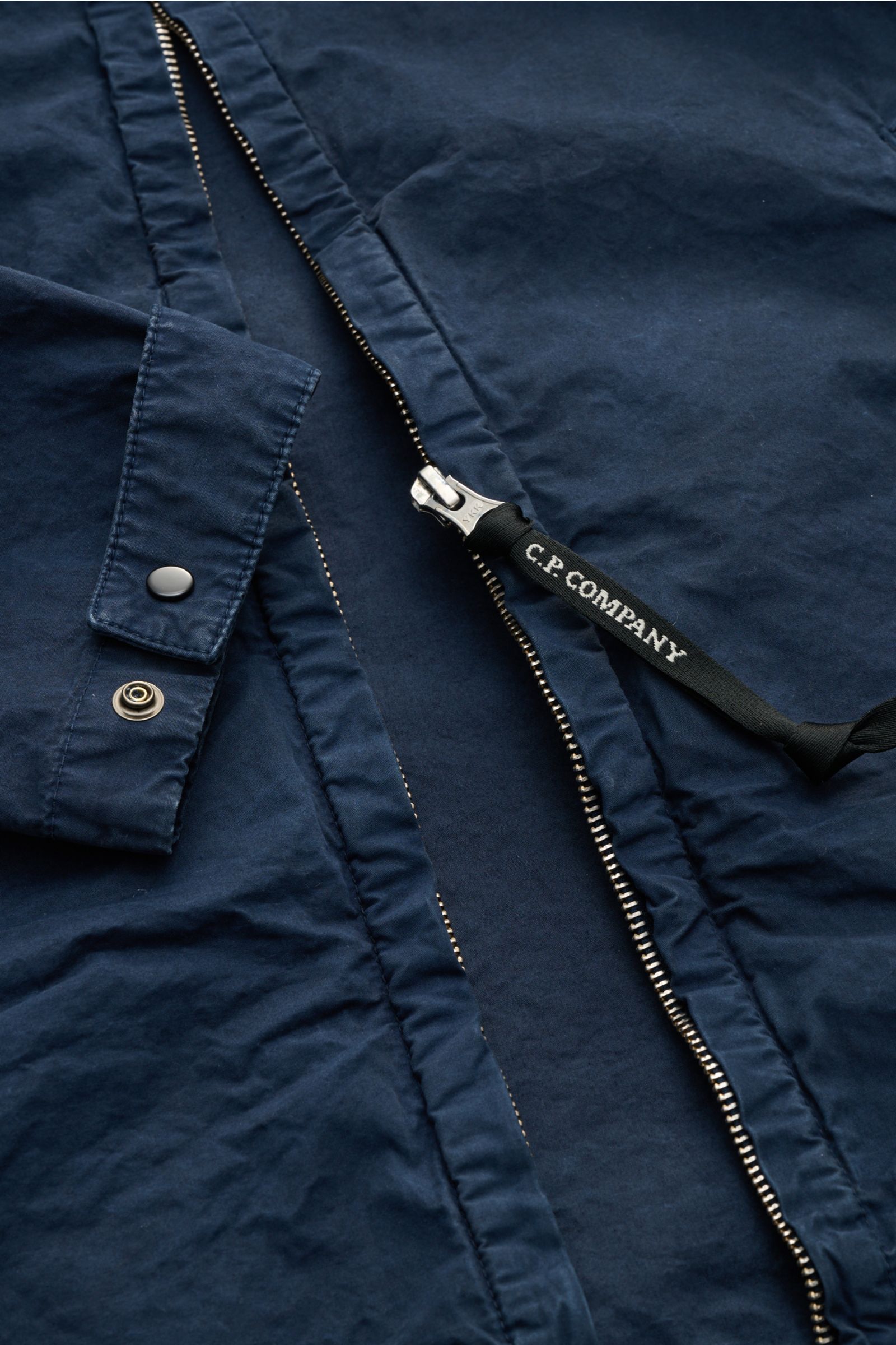 Nahaufnahme der C.P. Company Jacke navy, detailreich von oben fotografiert, zeigt Micro-Reps Baumwollstoff, Metall-Reißverschluss mit Logo und Manschette.