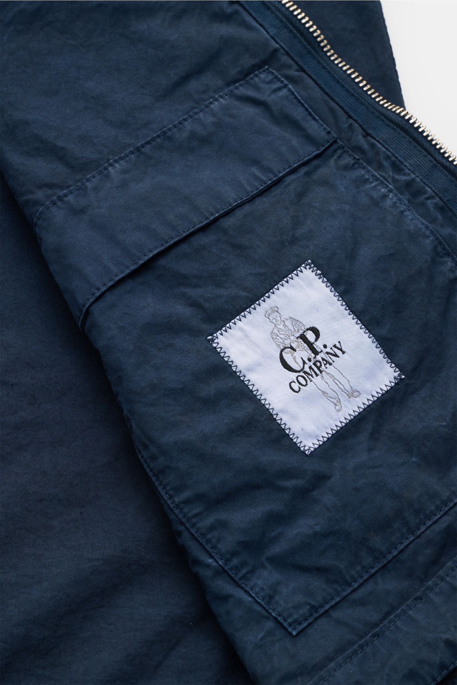 Close-up view of the C.P. Company Jacket navy sleeve showing the garment-dyed pure cotton fabric, metal zipper, and logo patch.