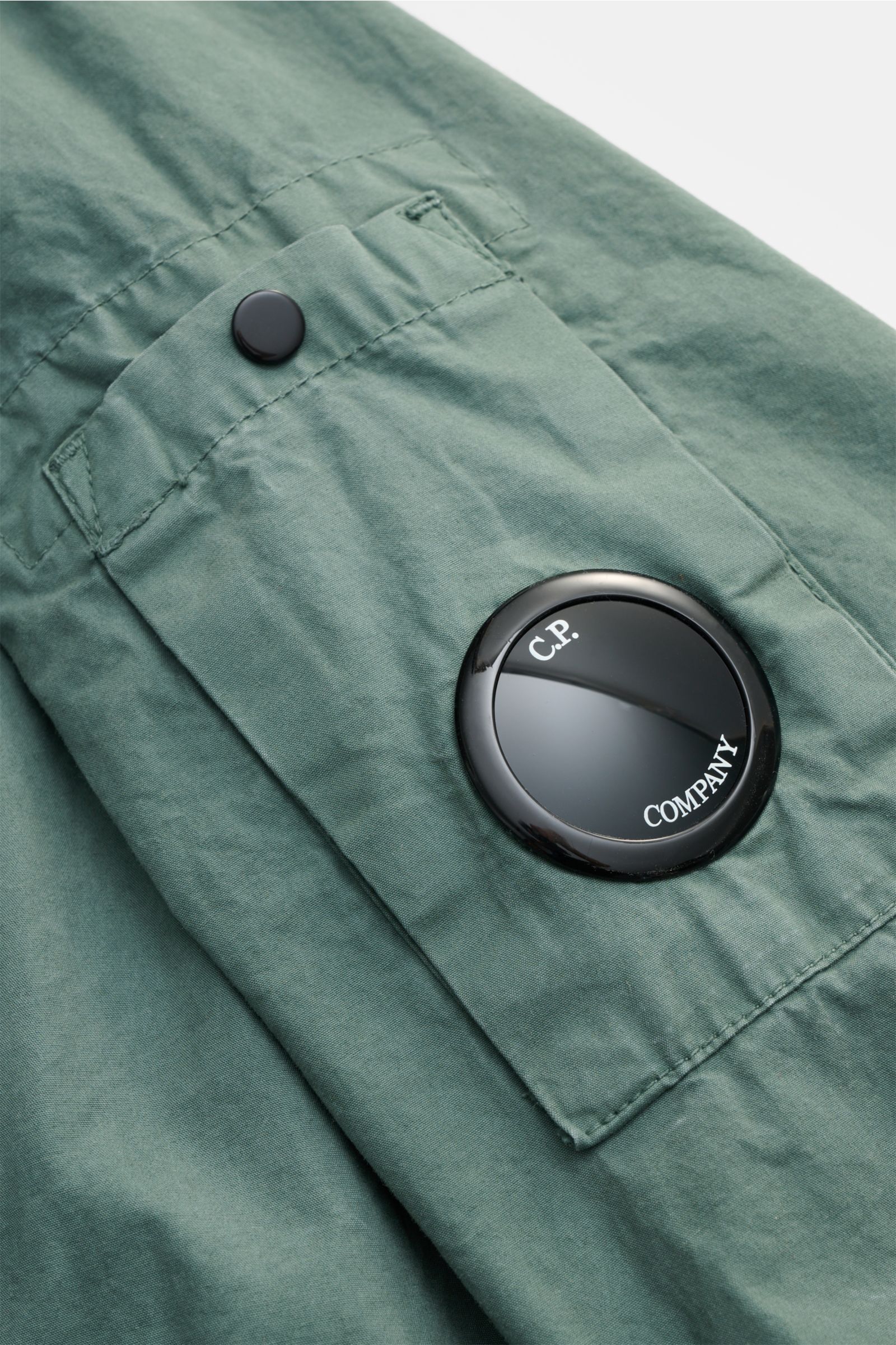 Close-up view of the C.P. Company Jacket sage pocket with black button and distinctive black lens logo, showing garment-dyed pure cotton fabric texture.