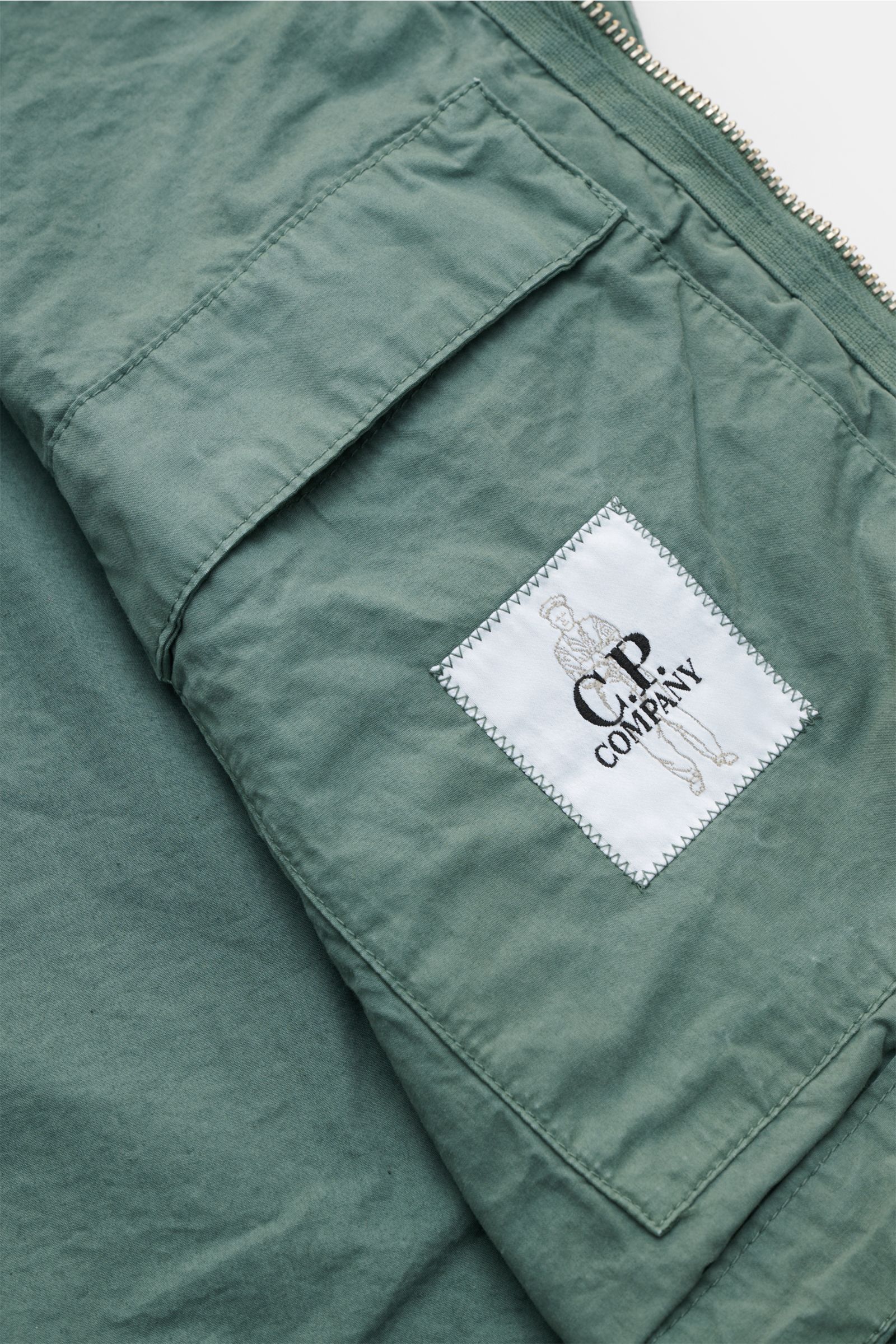 Close-up angled view of the C.P. Company Jacket sage showing the garment-dyed pure cotton fabric, sleeve pocket, and logo patch, emphasizing the lightweight and robust Micro-Reps textile.

Effortlessly urban: This new jacket with hood from 