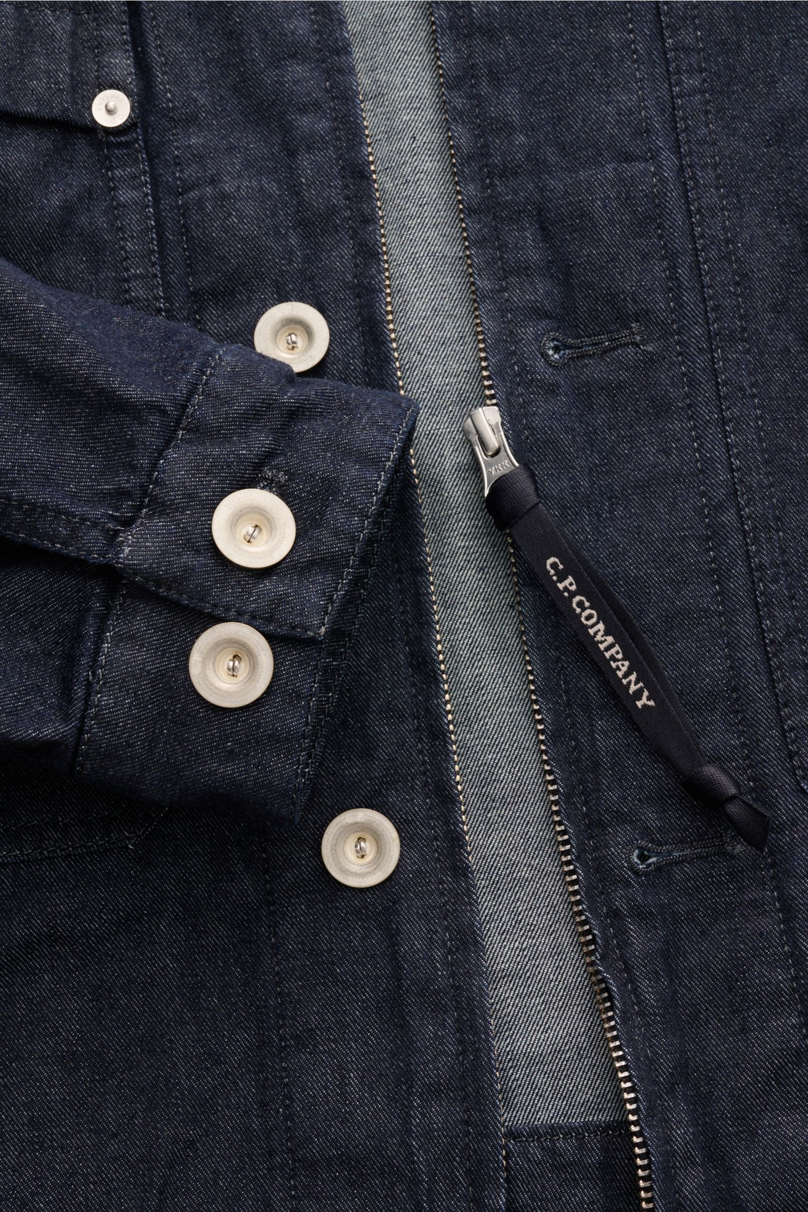 Close-up front view of the C.P. Company Jeansjacke navy showing slim fit, pure cotton fabric, buttoned sleeve cuffs, robust zipper with logo pull, and detailed stitching.